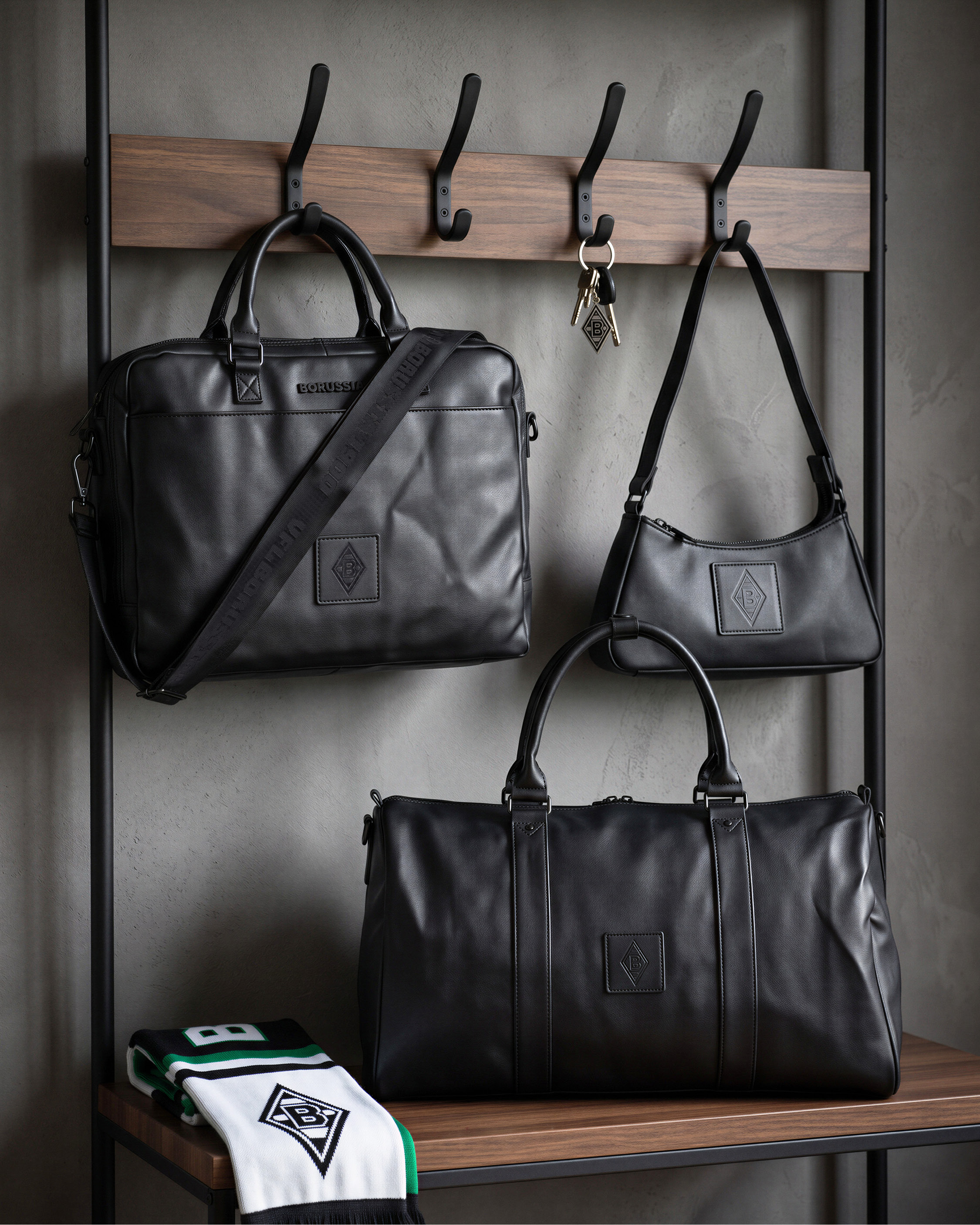 Three black leather bags hang on hooks and a shelf, alongside a white and green scarf and keys on a wooden and metal rack.