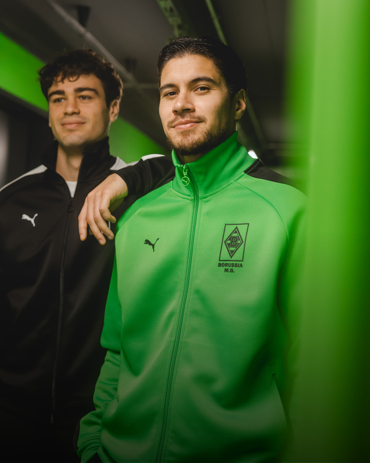 Two men wearing Borussia Mönchengladbach tracksuits, one in green and one in black, posing confidently indoors against a green-lit background.