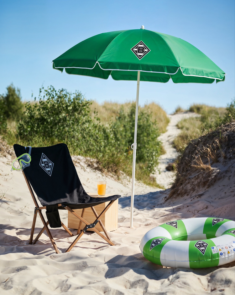 Beach scene with a green umbrella, black chair, snorkel, juice on a wooden box, and a green-and-white inflatable ring on sandy path with shrubs.