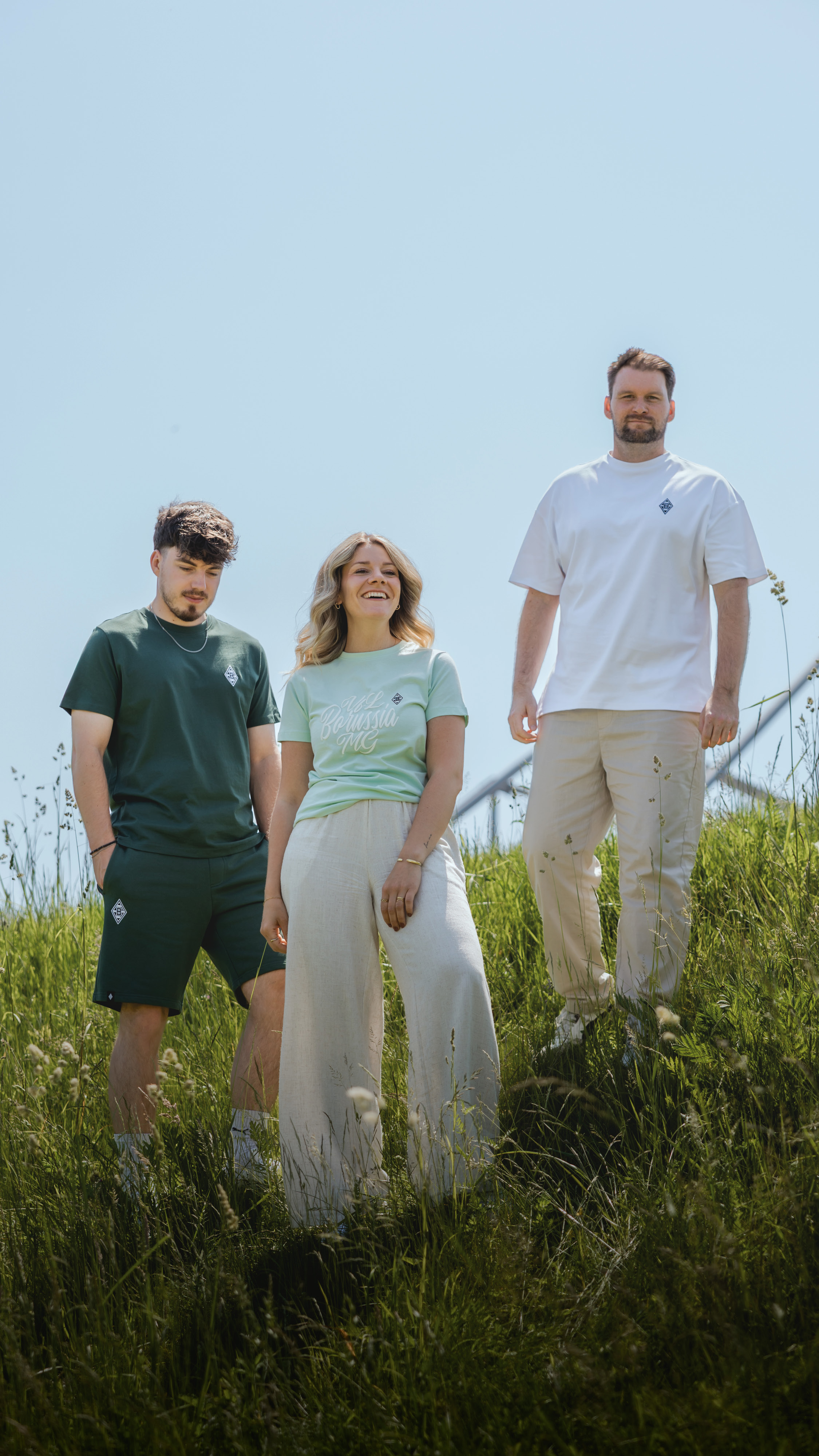 Three people stand on a grassy hill under a clear sky, dressed casually in shorts, trousers, and t-shirts, smiling and enjoying the sunny day.