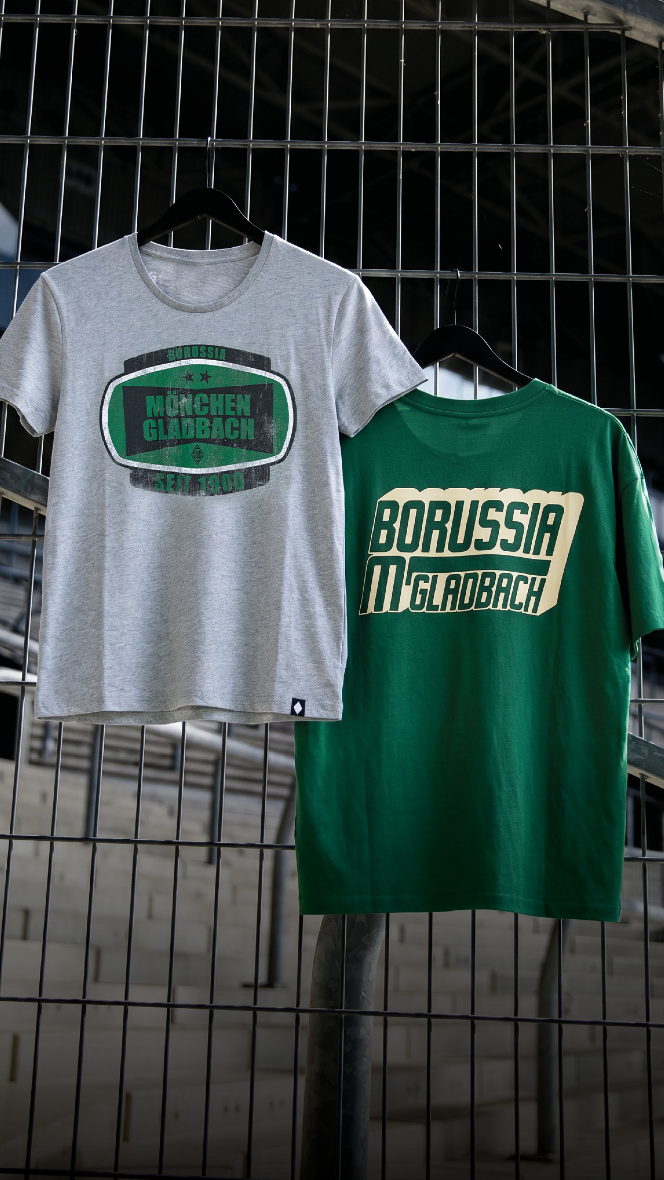 Two Borussia Mönchengladbach t-shirts, one gray and one green, hang on a fence in a stadium setting.