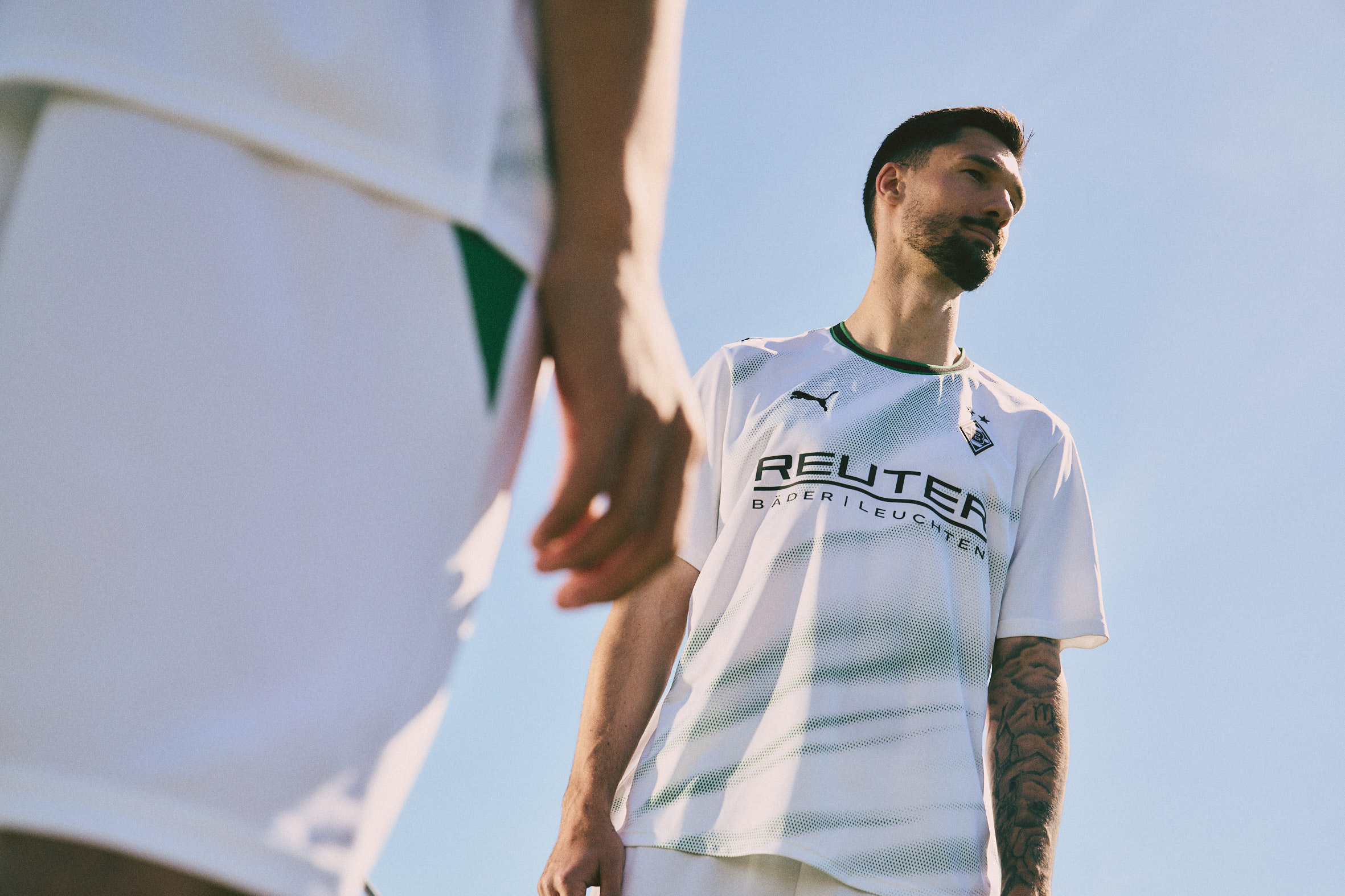 Two soccer players in white jerseys, one in the foreground from the waist down, another looking upward under a clear blue sky.