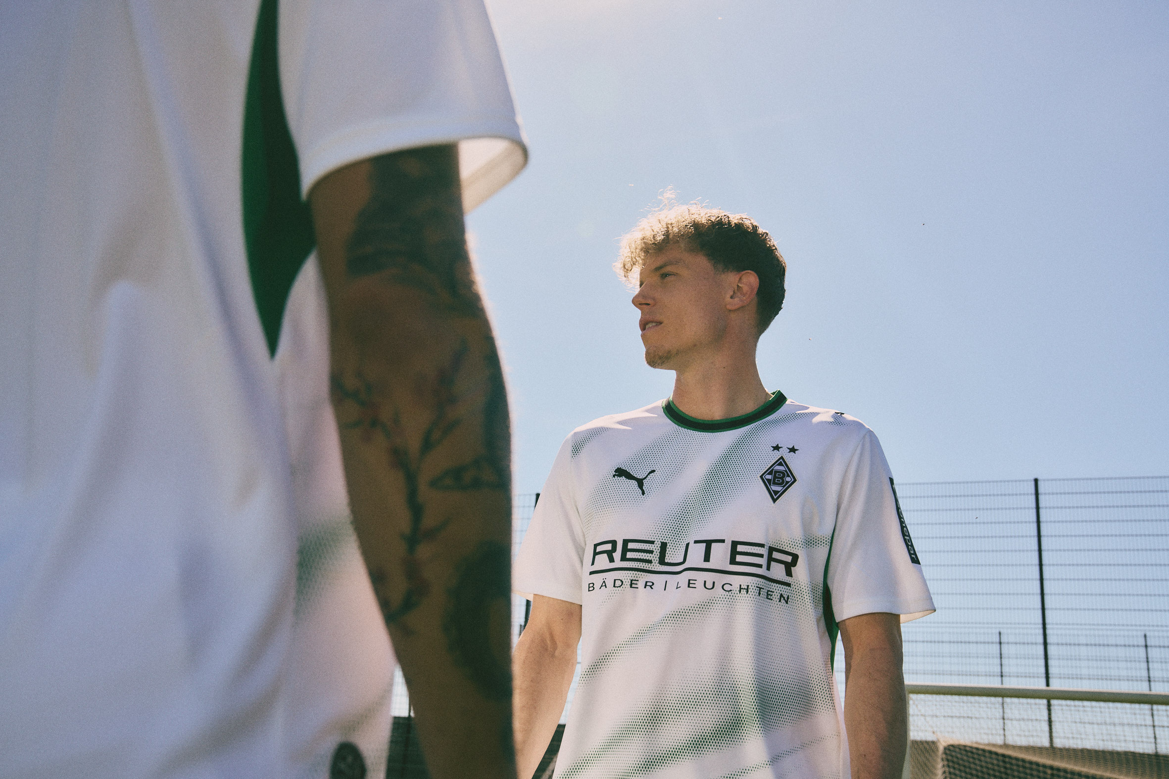 Two men wearing soccer jerseys stand on a field under a clear blue sky, one tattooed arm visible.