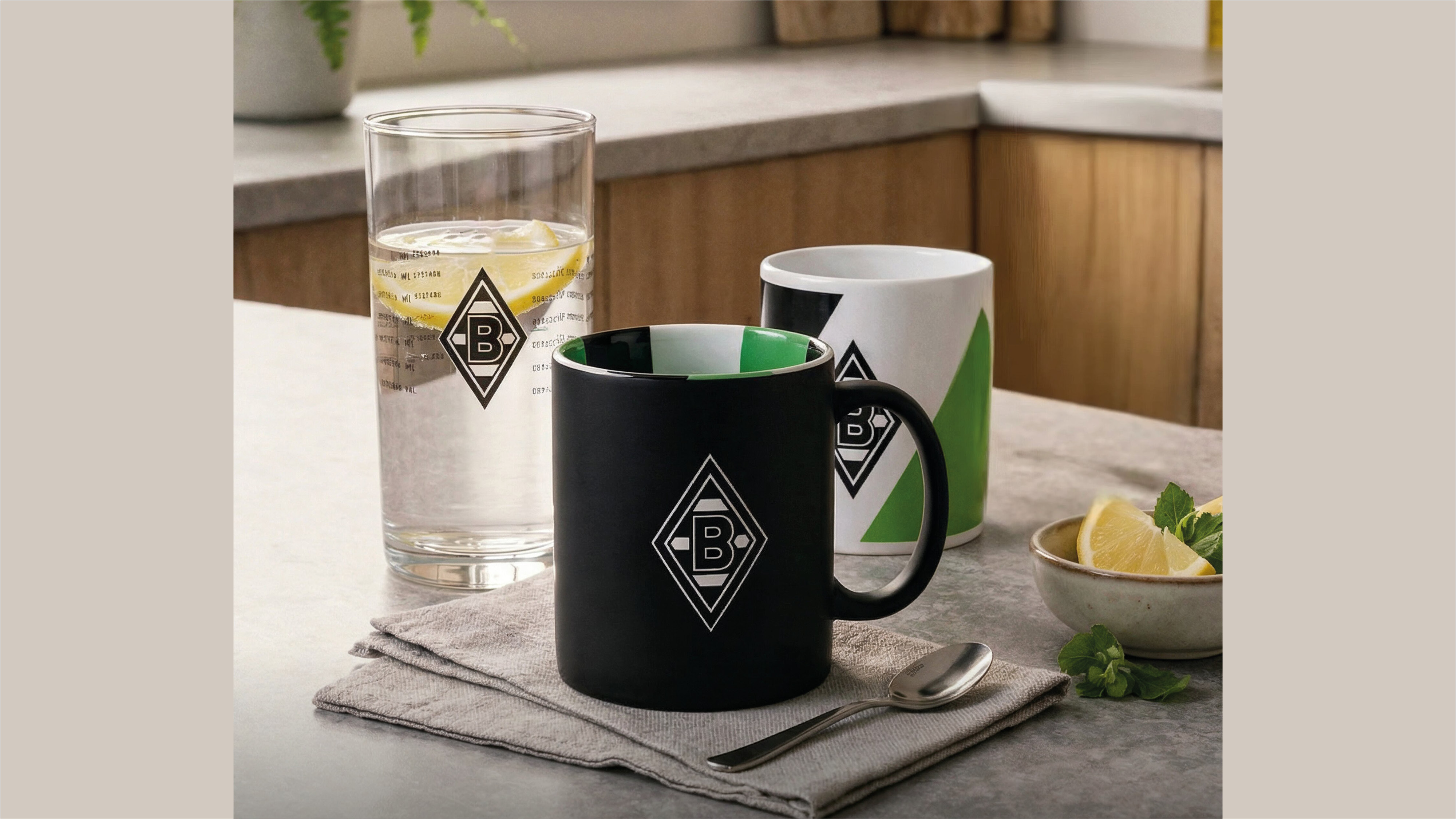 A black mug, white mug, and glass with lemon slices sit on a white napkin in a kitchen, each featuring a logo with the letter "B".