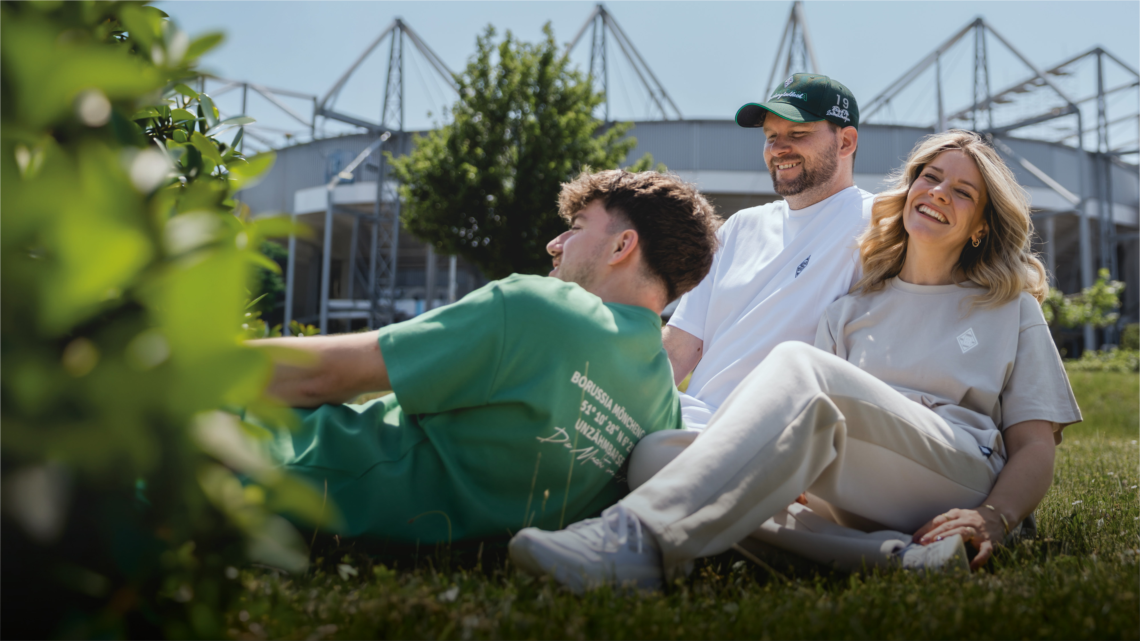 Three people relaxing on grass near a stadium, smiling and enjoying a sunny day.