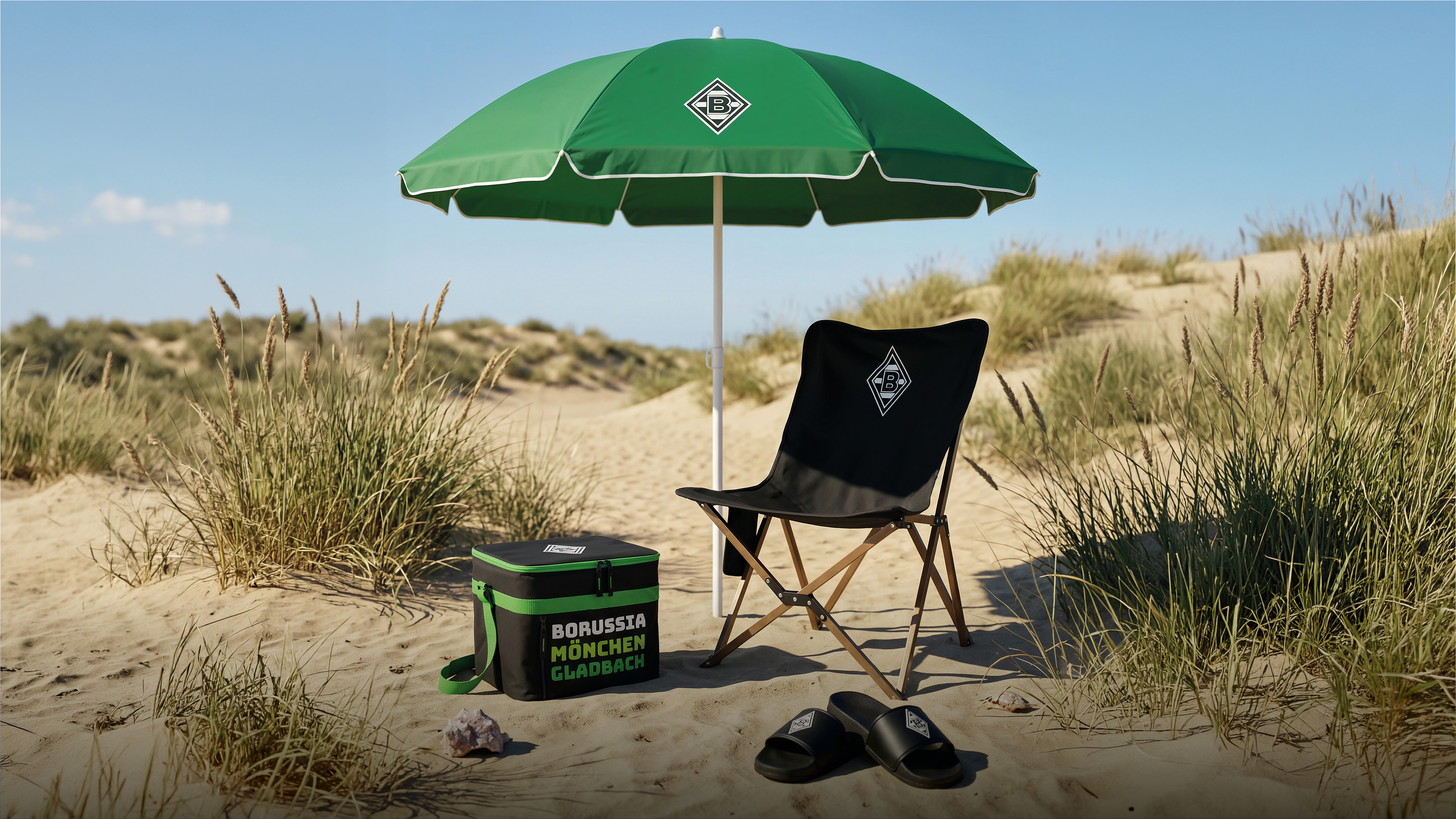 Green umbrella, black chair, cooler, and sandals with "Borussia Mönchengladbach" logos on a sandy beach with grass dunes under a clear blue sky.