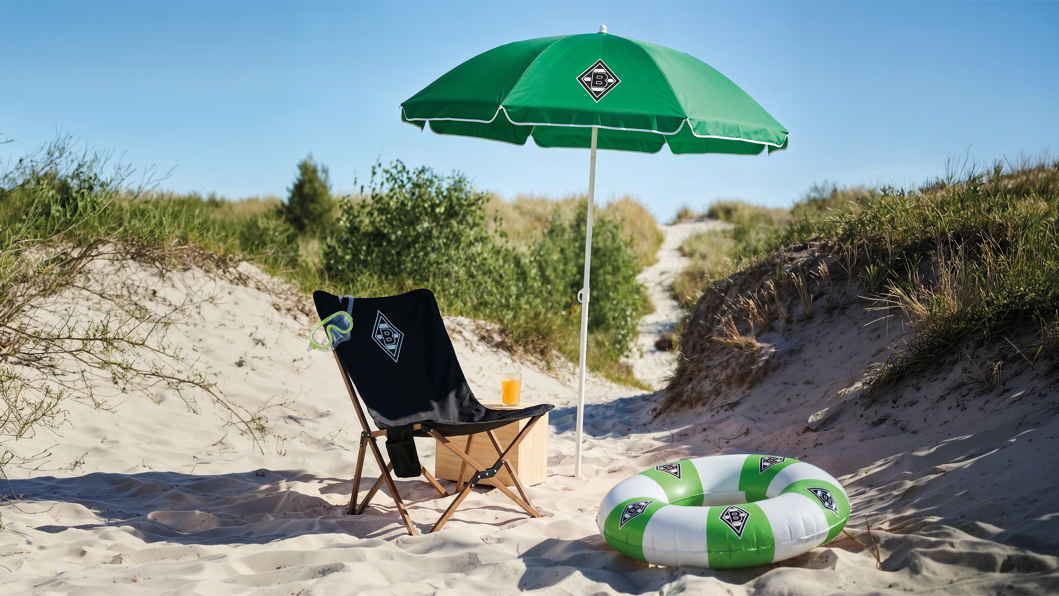 Beach scene with a green umbrella, black chair, float ring, and a drink on sand dunes under a clear blue sky.