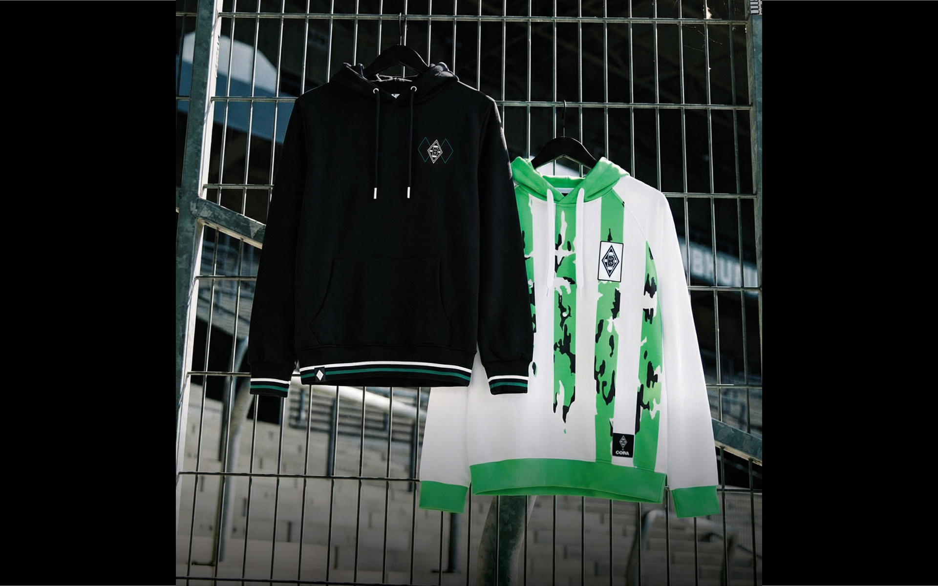 Two hoodies hang on a metal fence; one is black with a logo, and the other is white with green accents and graphic design.