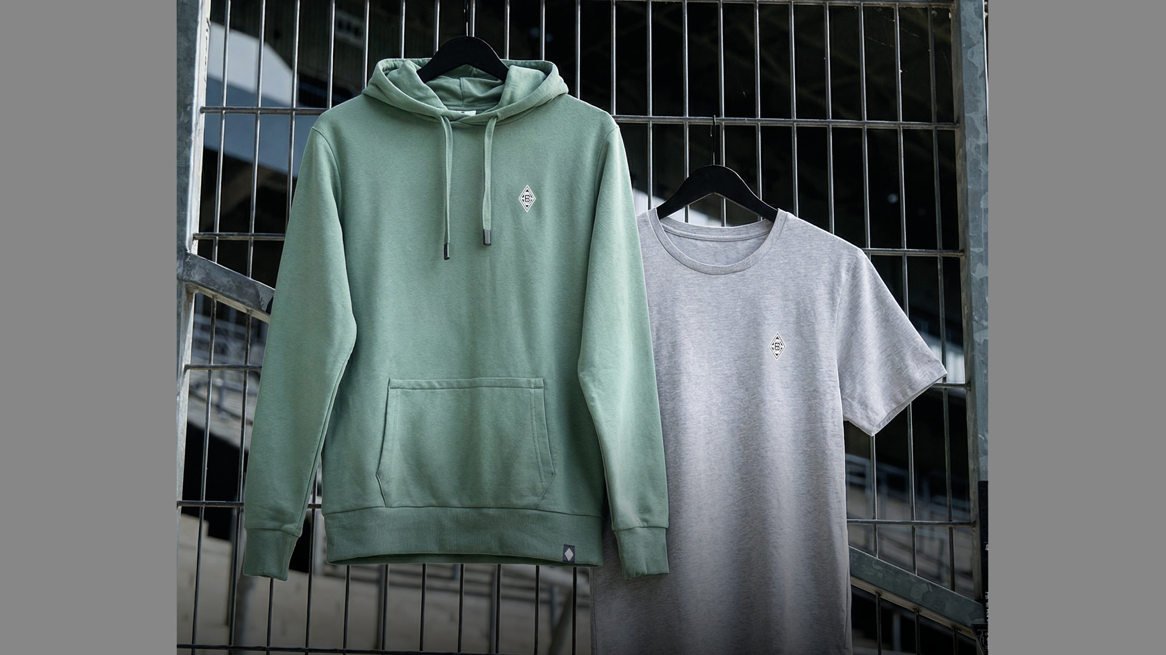 Green hoodie and gray T-shirt on hangers against a metal grate background. Each garment has a small embroidered emblem.