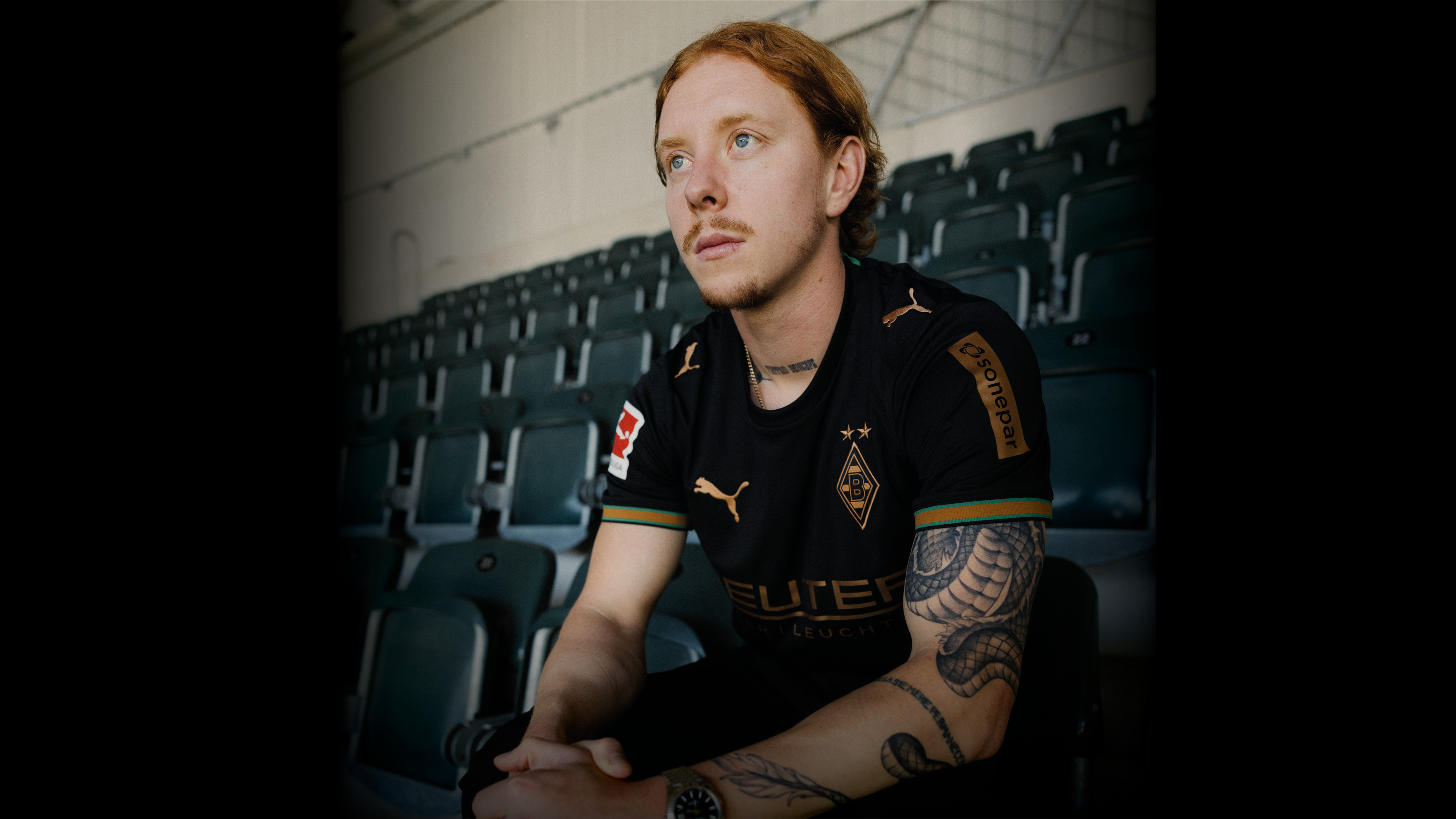 A man with tattoos sits in a stadium, wearing a black Borussia Mönchengladbach jersey.