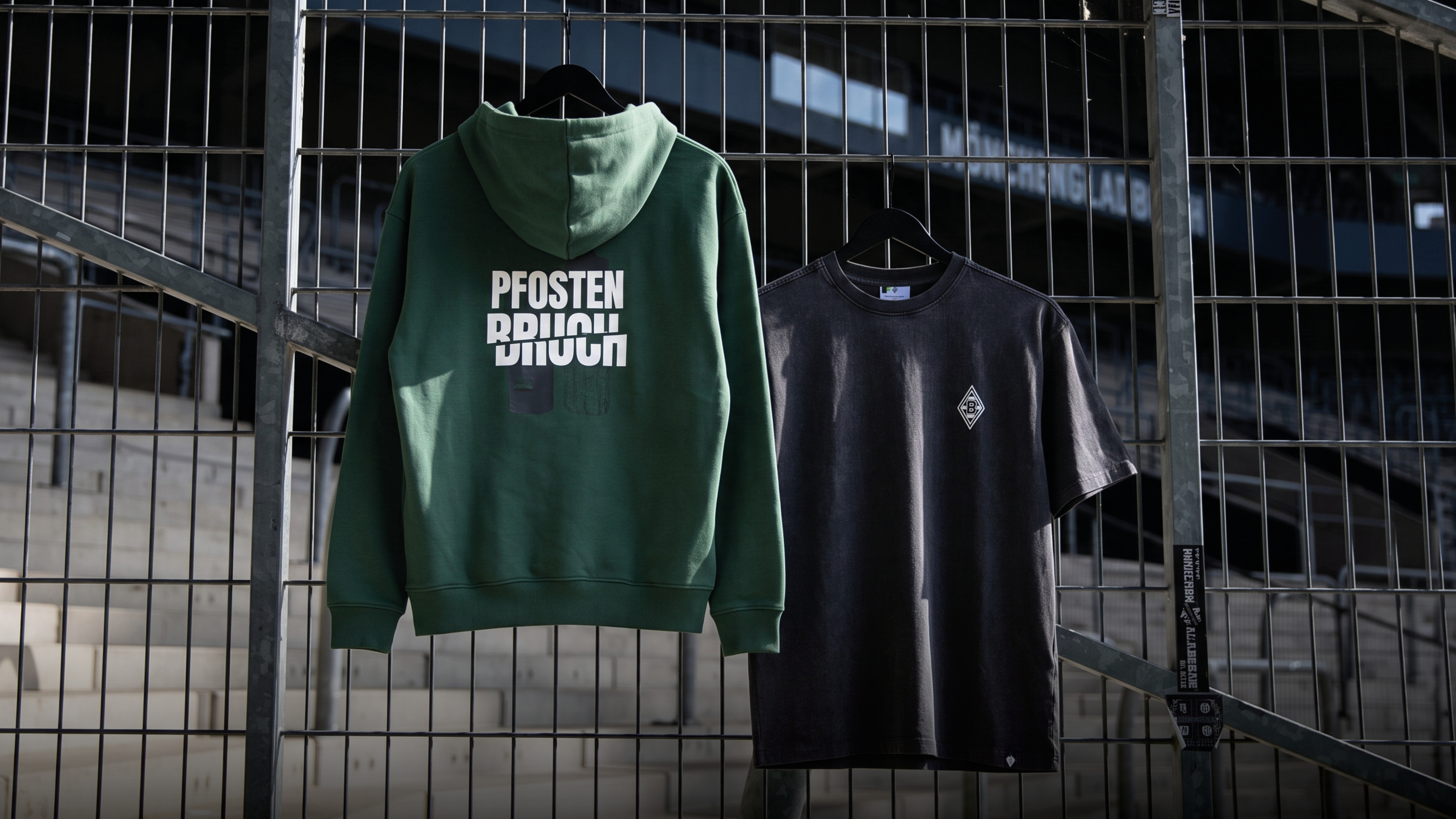 A green hoodie and a dark T-shirt hang on a metal fence, with bleachers visible in the background.