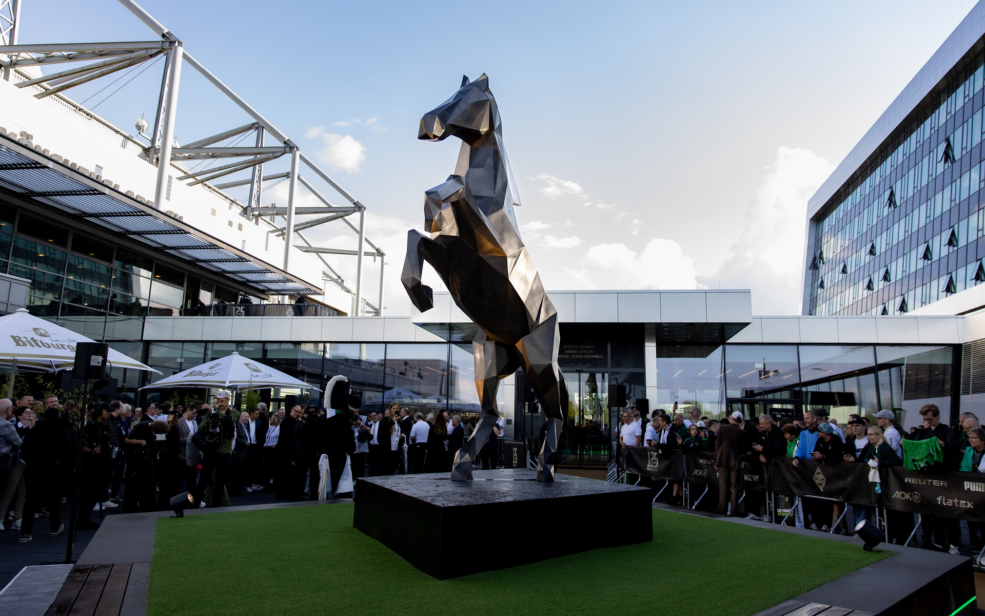 A large metallic horse sculpture stands on a platform, surrounded by a crowd, with modern buildings in the background.