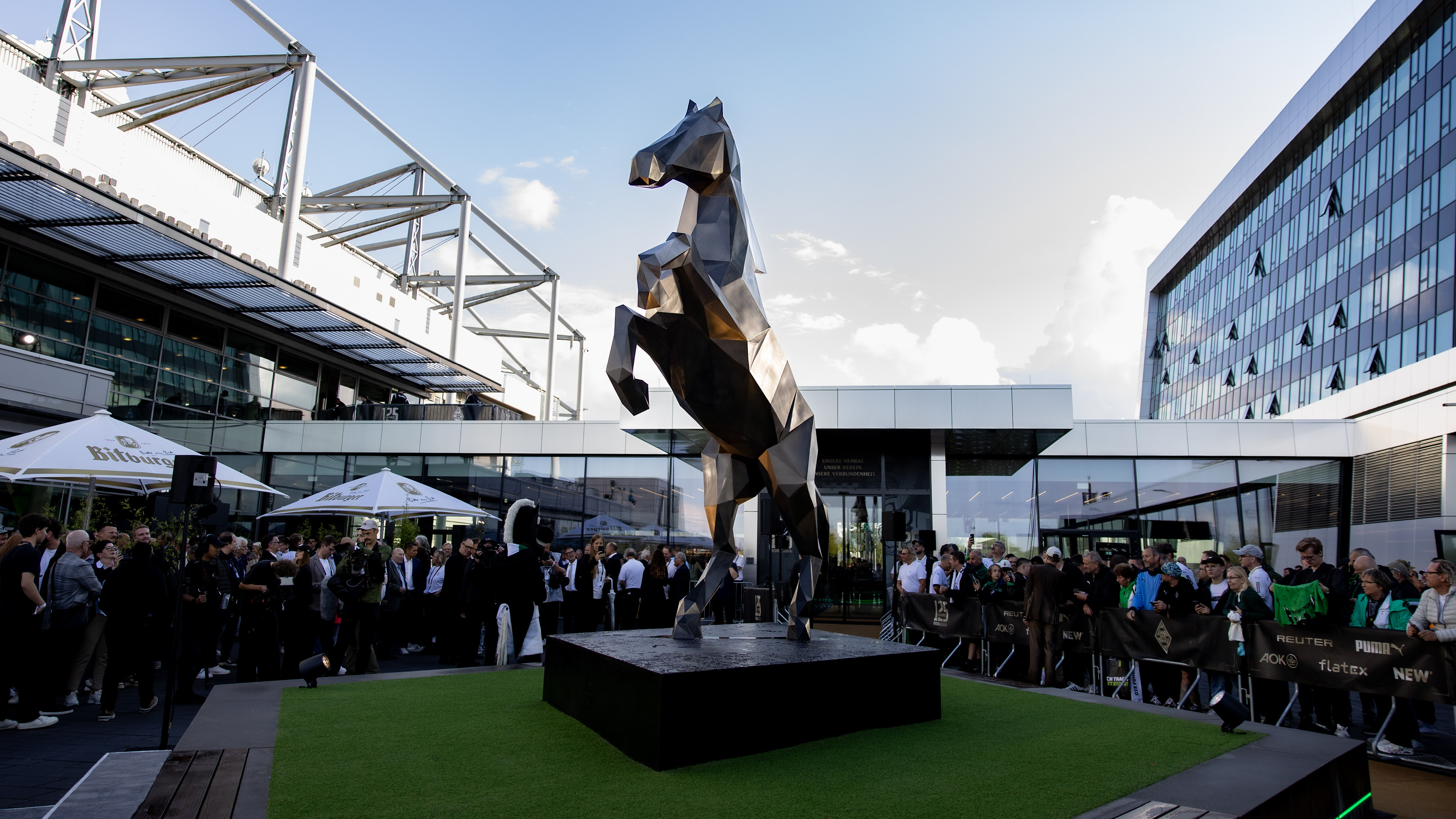 A large metallic horse sculpture stands on a platform, surrounded by a crowd, with modern buildings in the background.