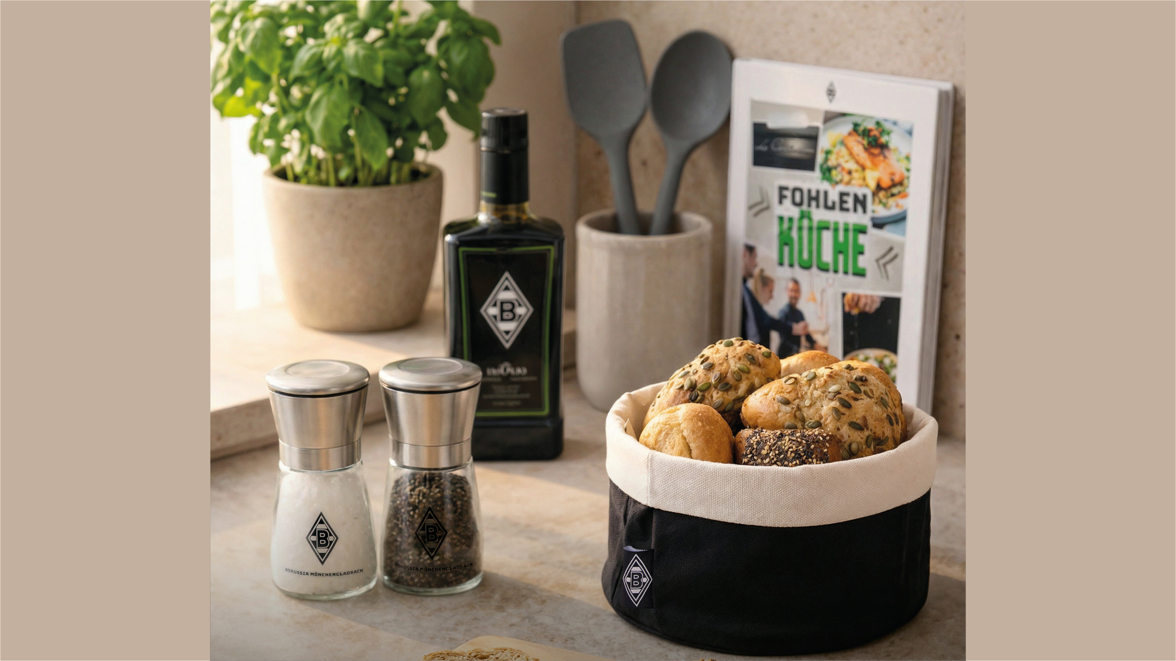 A kitchen countertop with herb plant, olive oil bottle, cookbook, utensils, salt and pepper shakers, and a basket of mixed bread rolls.