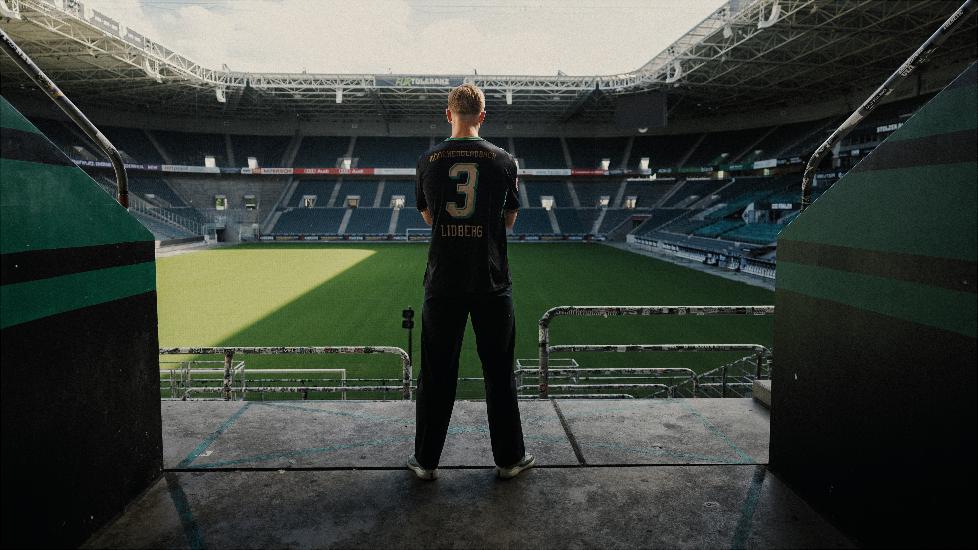 A person in a black jersey stands in a soccer stadium tunnel, overlooking the green field and empty stands.