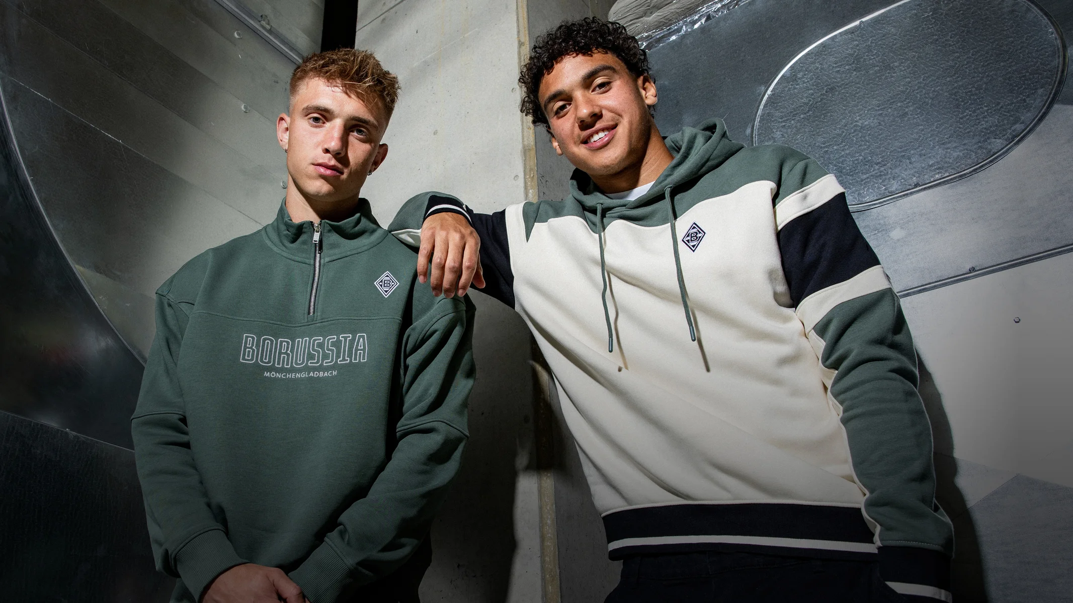 Two men in casual Borussia-themed sweatshirts pose confidently against an industrial backdrop, featuring metallic and concrete elements.