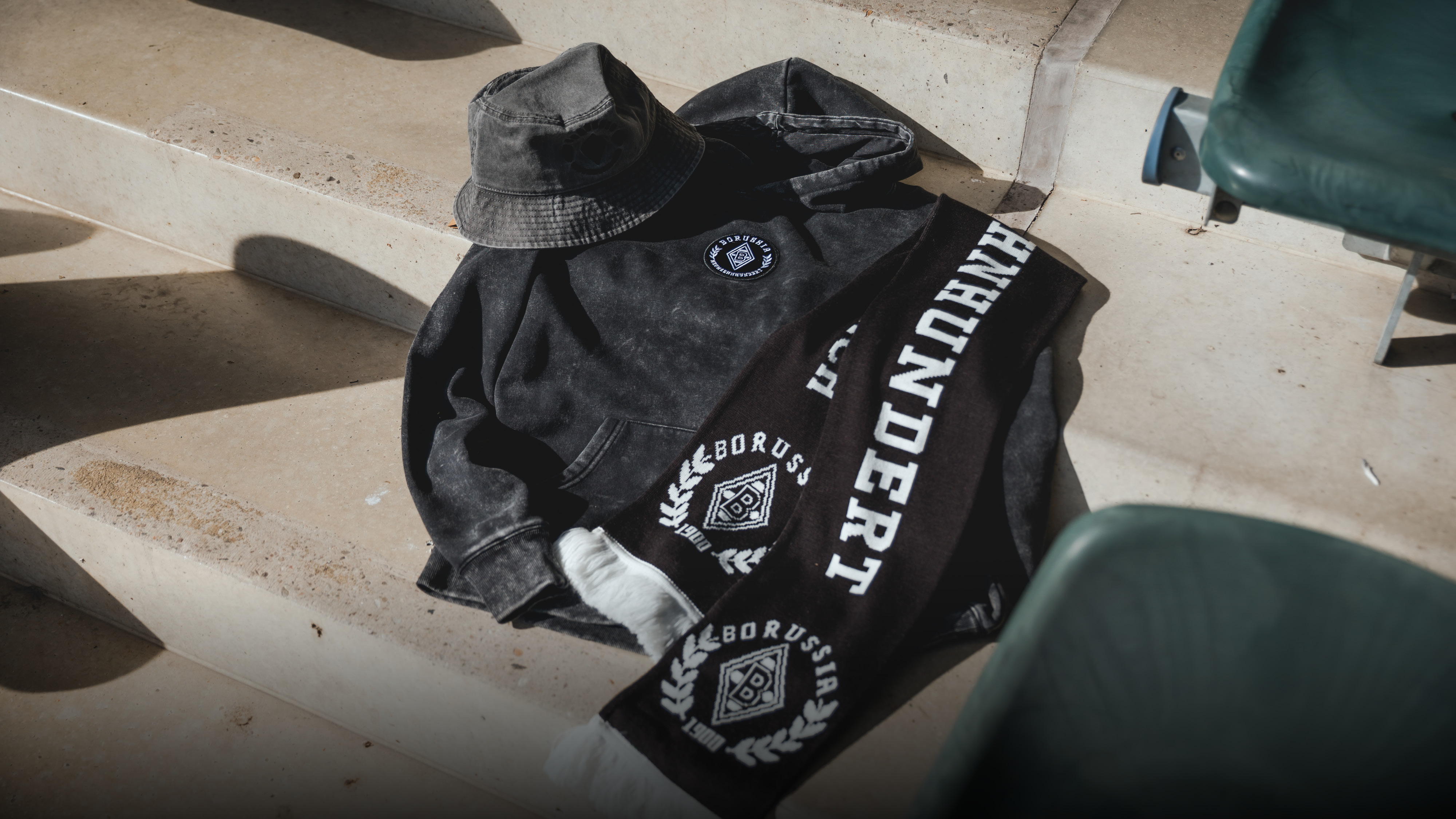 Black bucket hat, hoodie, and scarf with "Borussia" logo and "HUNDERT" text, laid on stone steps with sunlight and shadows.