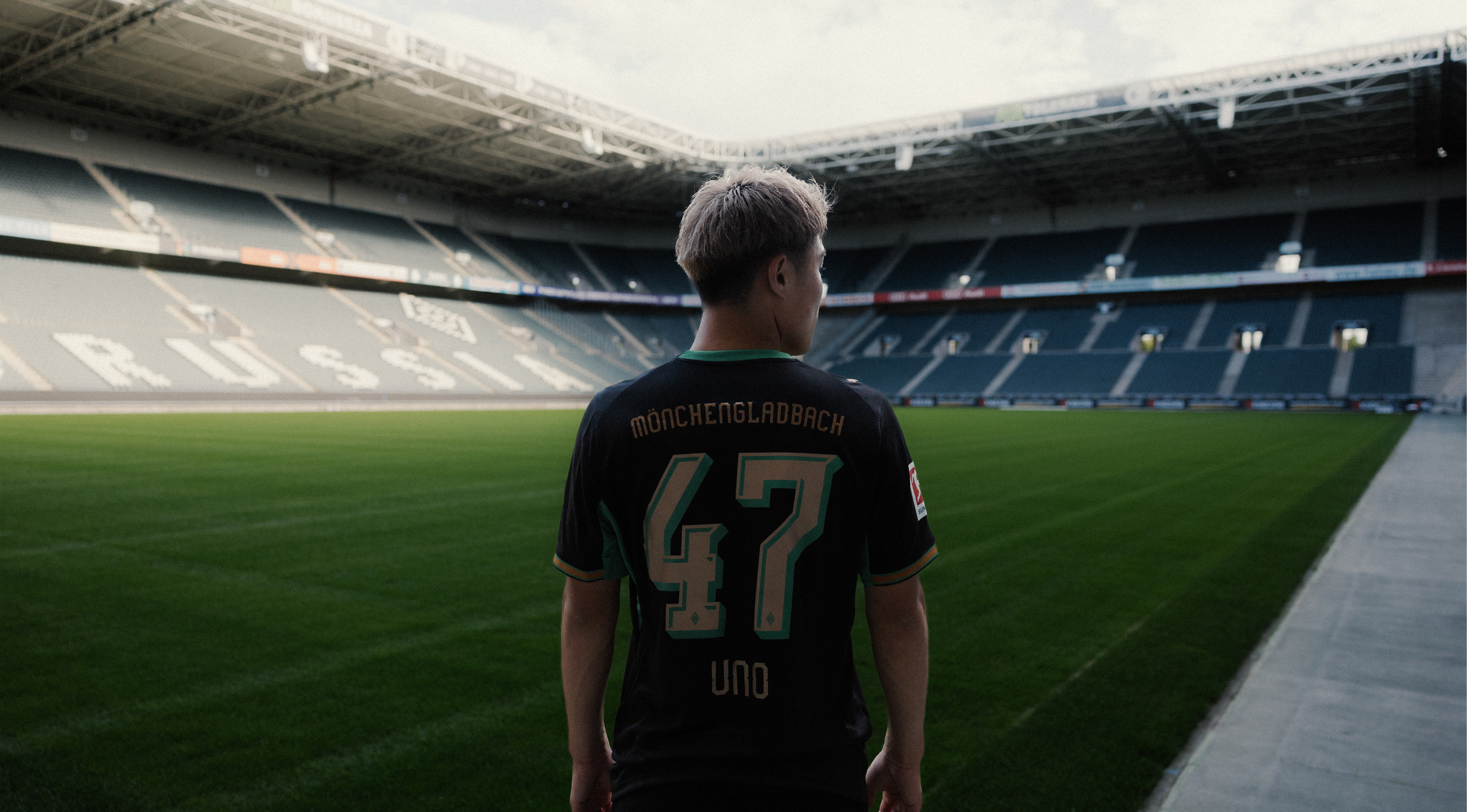 A person in a Mönchengladbach jersey with the number 47 stands on a soccer field inside a large, empty stadium.