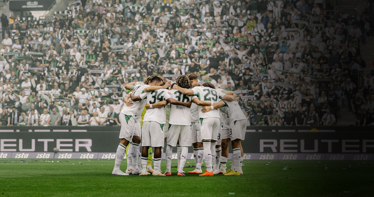 Soccer team huddle on the field, players wearing white jerseys, with a crowd of fans in the background.
