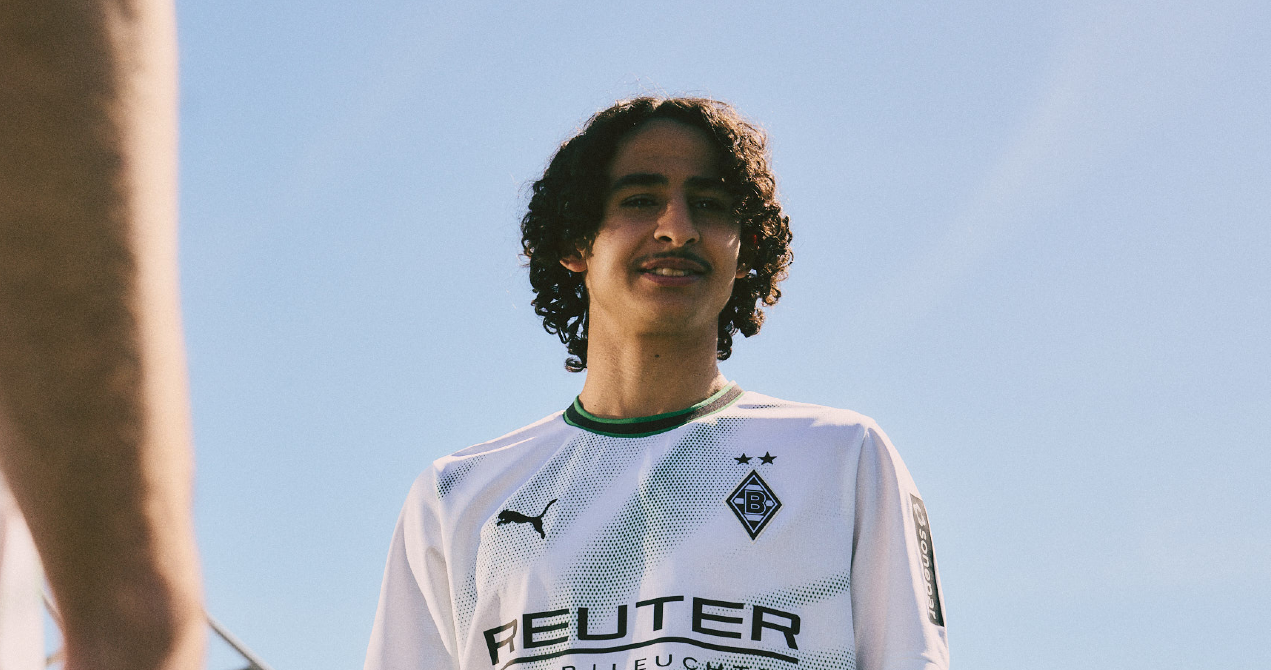 Individual with curly hair wearing a Borussia Mönchengladbach jersey looks up under a clear blue sky.