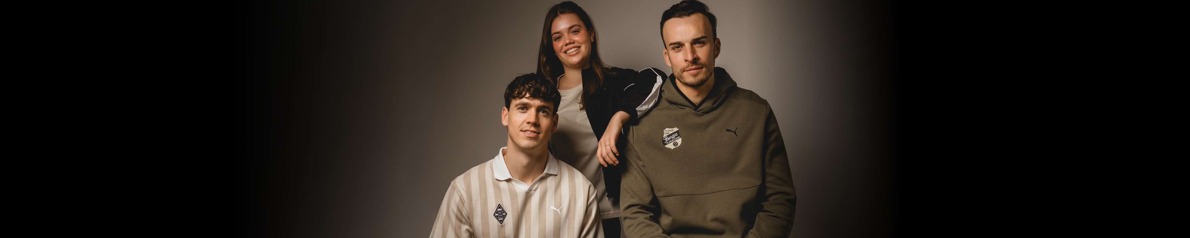 Three people posing against a neutral background, two seated and one standing behind them, all casually dressed and smiling slightly.