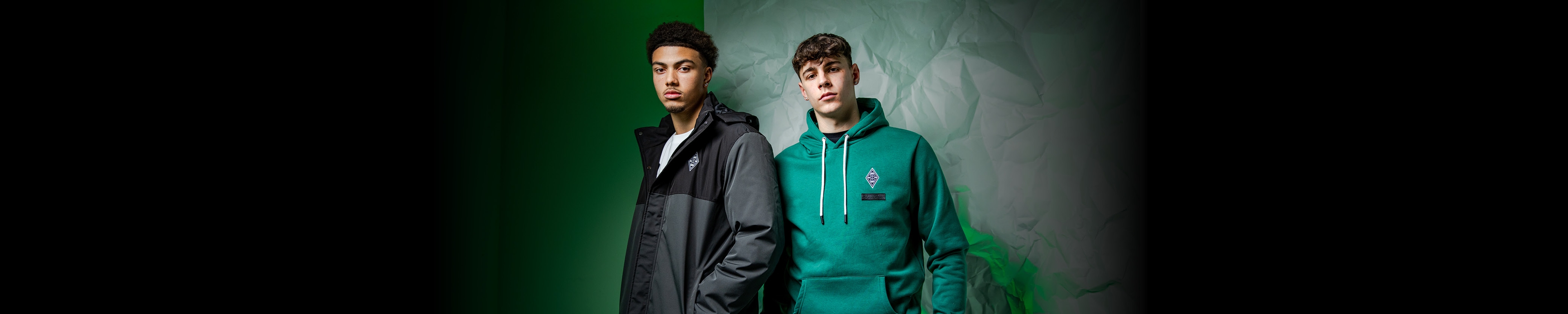 Two men in casual wear; one in a dark coat, the other in a green hoodie with a logo, against a green and gray textured background.