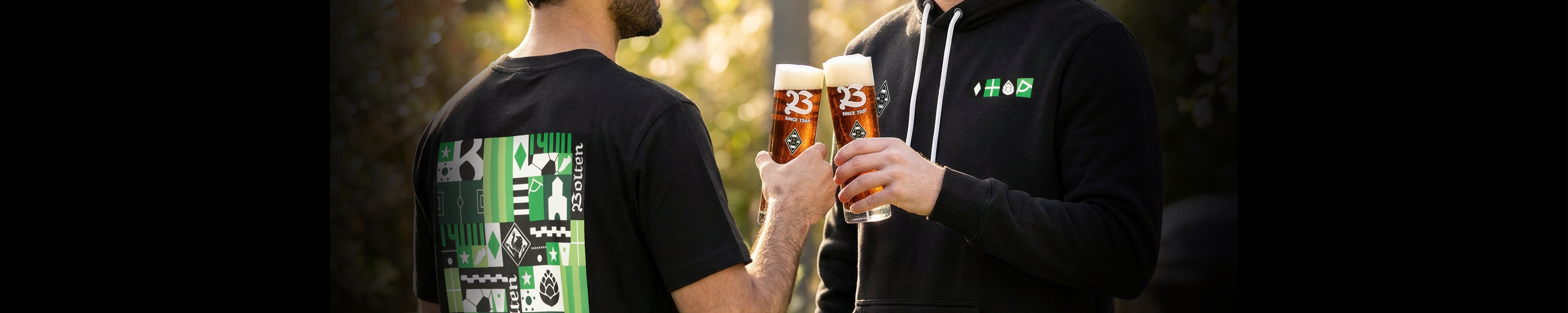 Two people clink glasses filled with amber beer, wearing black shirts with green and white designs, outdoors in soft lighting.