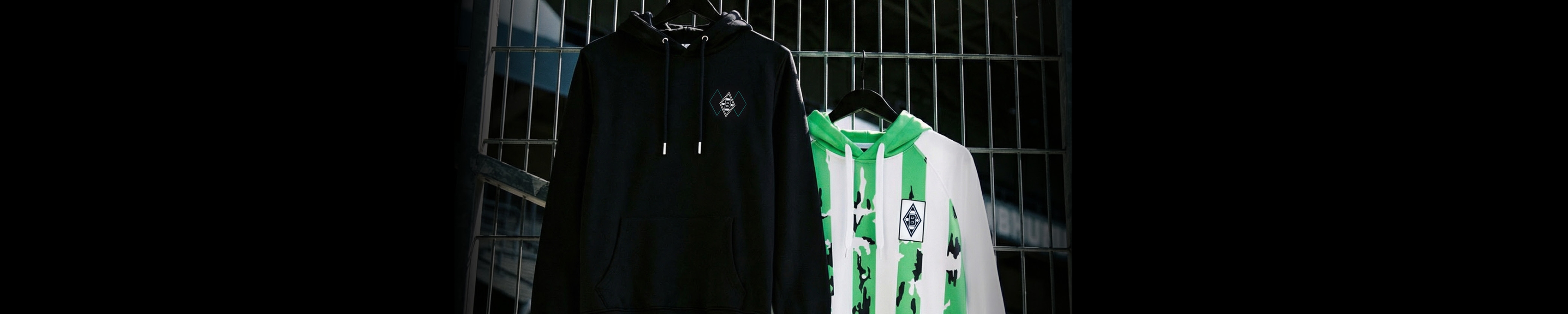 Two soccer jerseys hang on a rack: one is black with a logo, the other is white with green stripes and a crest.
