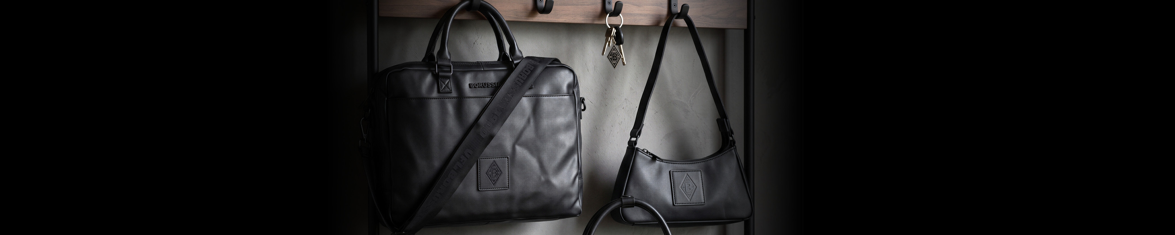Two black leather bags hanging on wooden hooks, one larger with a handle and shoulder strap, the other smaller with a curved shape.