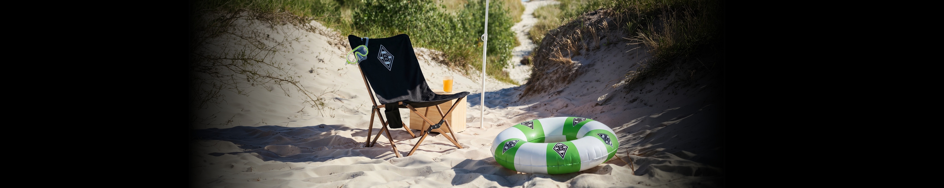 A sunny beach scene with a folding chair, a green and white inflatable ring, and a sandy path leading through dunes with grass.
