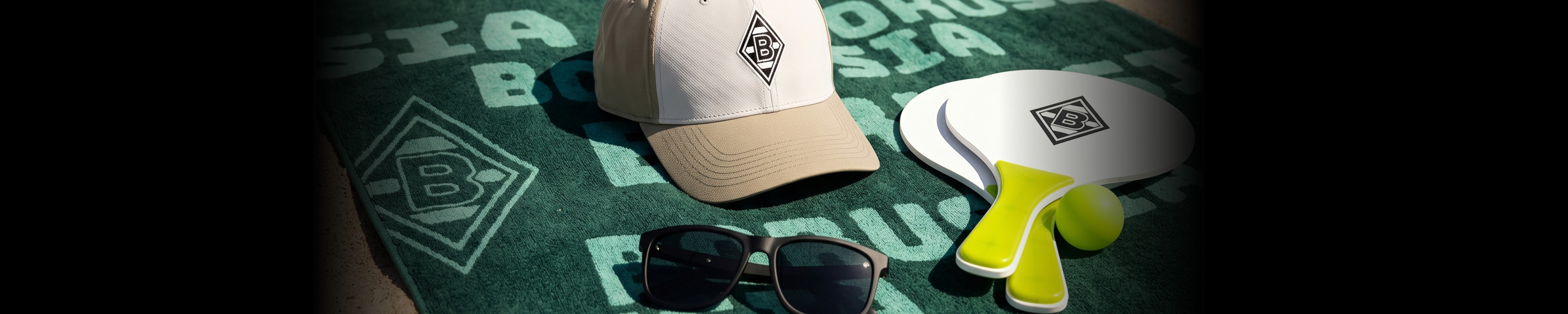 A cap, sunglasses, and two beach rackets lie on a green Borussia towel with logos, set against a dark background.