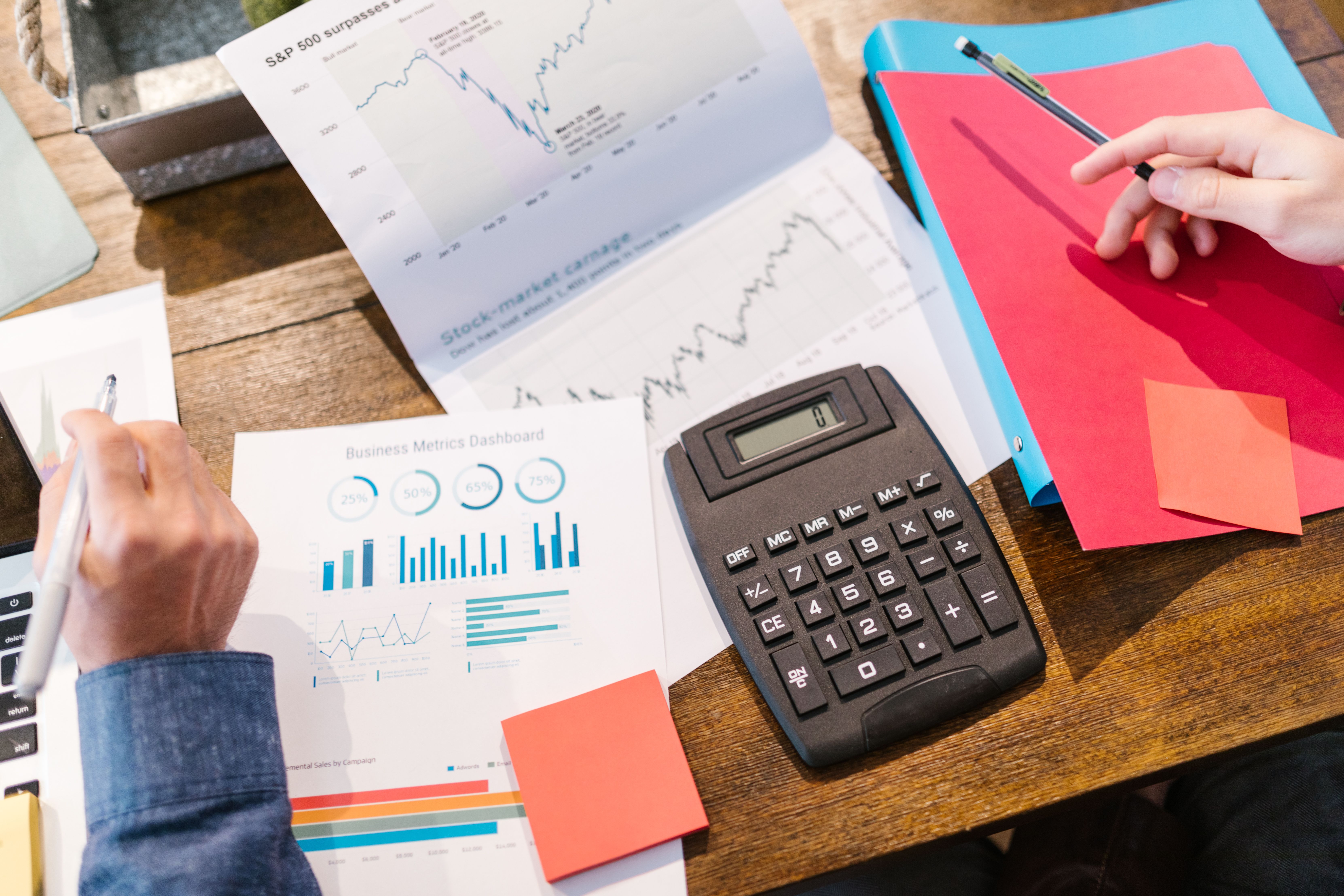 Hands holding pens over business graphs, charts, a calculator, and colored folders on a wooden desk.