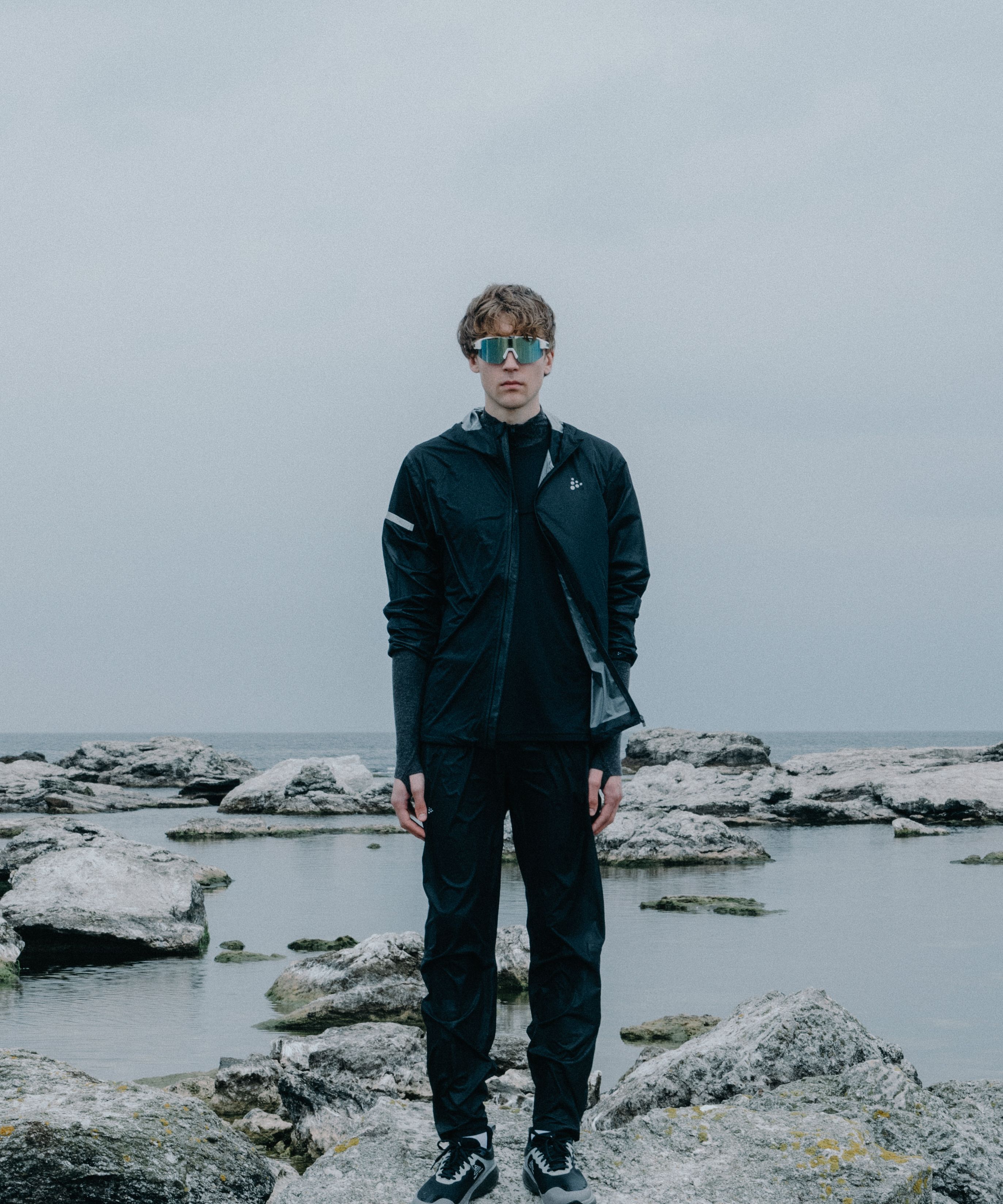 A person in dark athletic wear and reflective sunglasses stands on rocky terrain by the sea under a cloudy sky.
