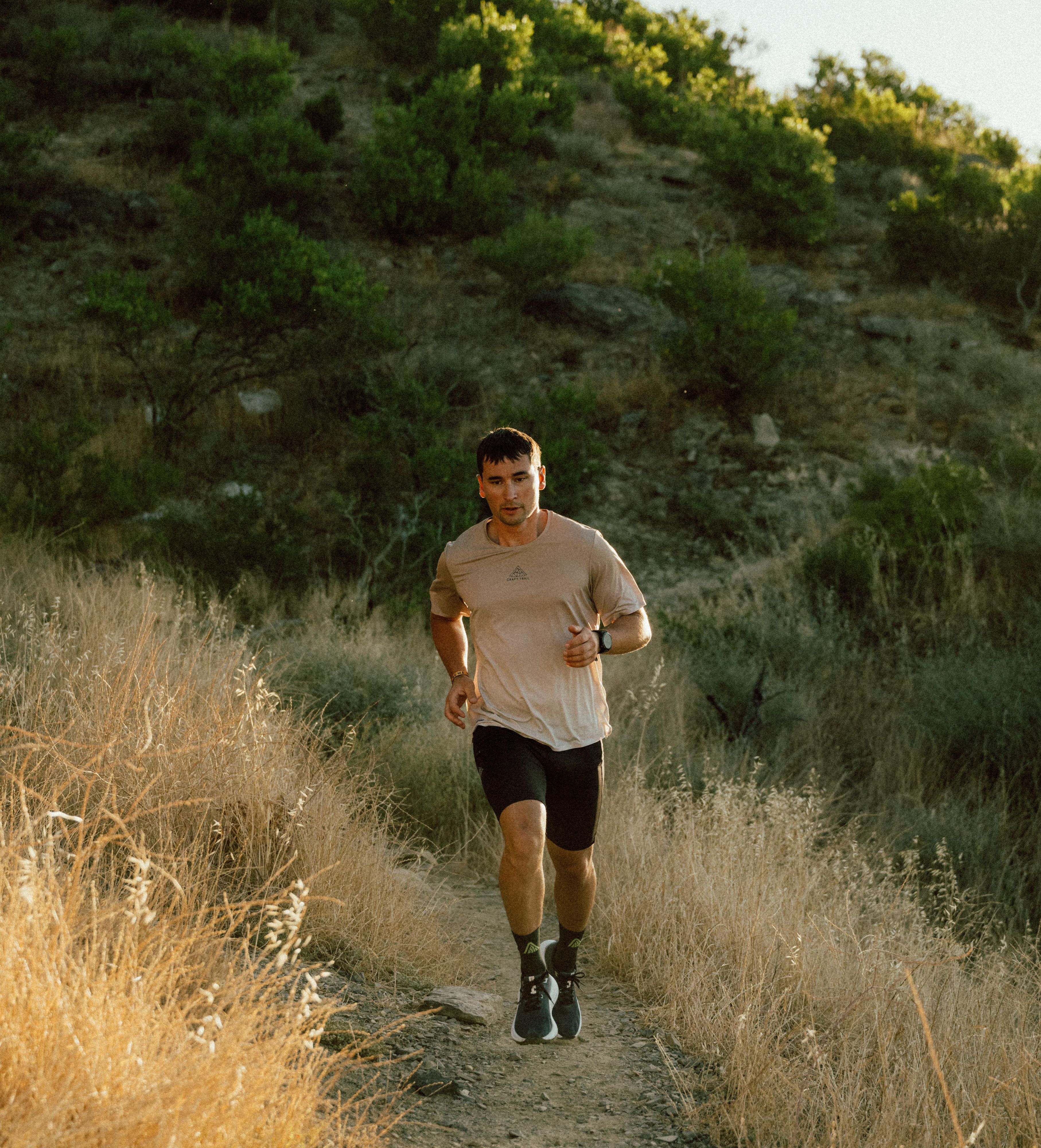 A man in athletic wear runs along a narrow dirt trail surrounded by dry grass and greenery on a hillside under a clear sky.