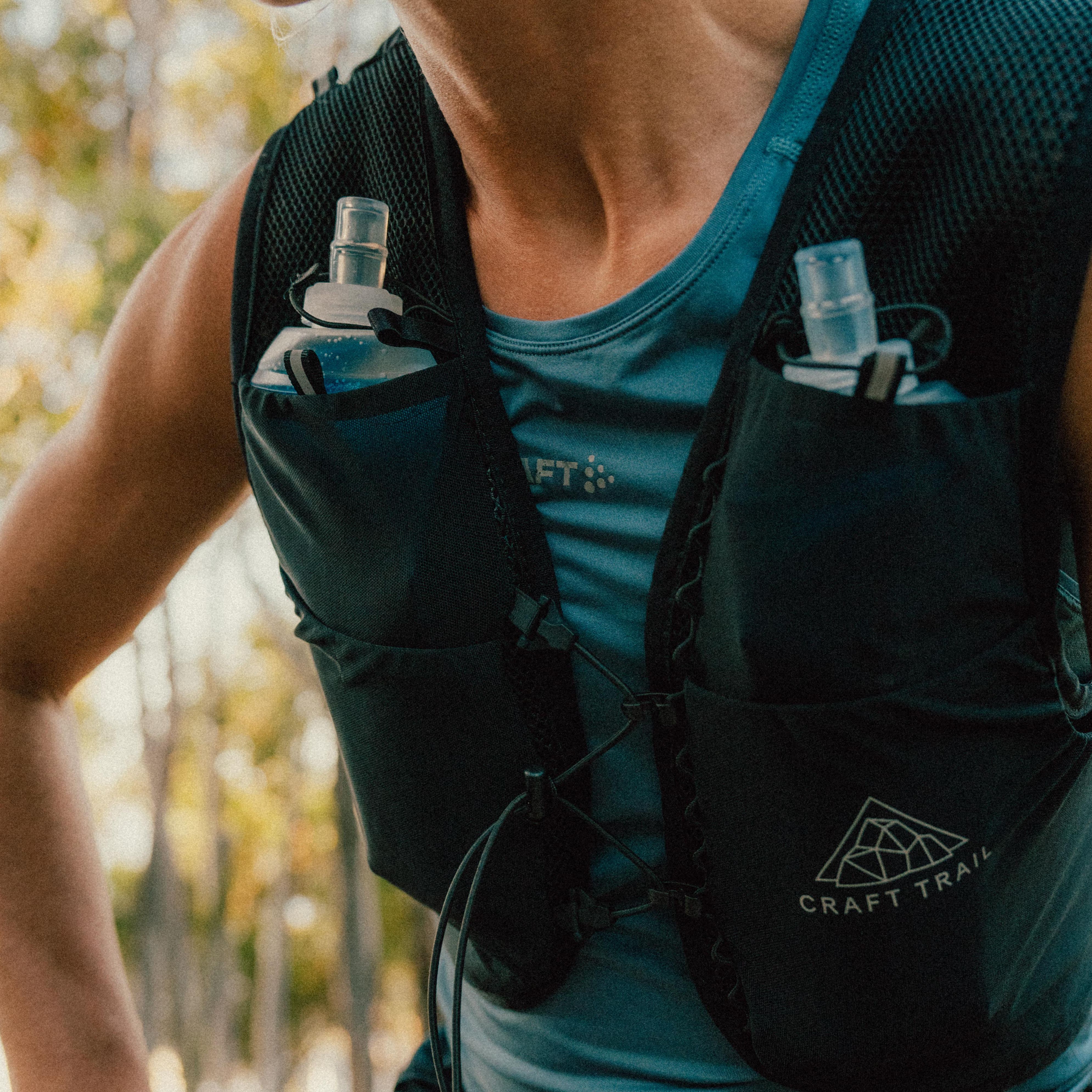 Person wearing a blue running vest with water bottles, standing outdoors. Trees are blurred in the background.