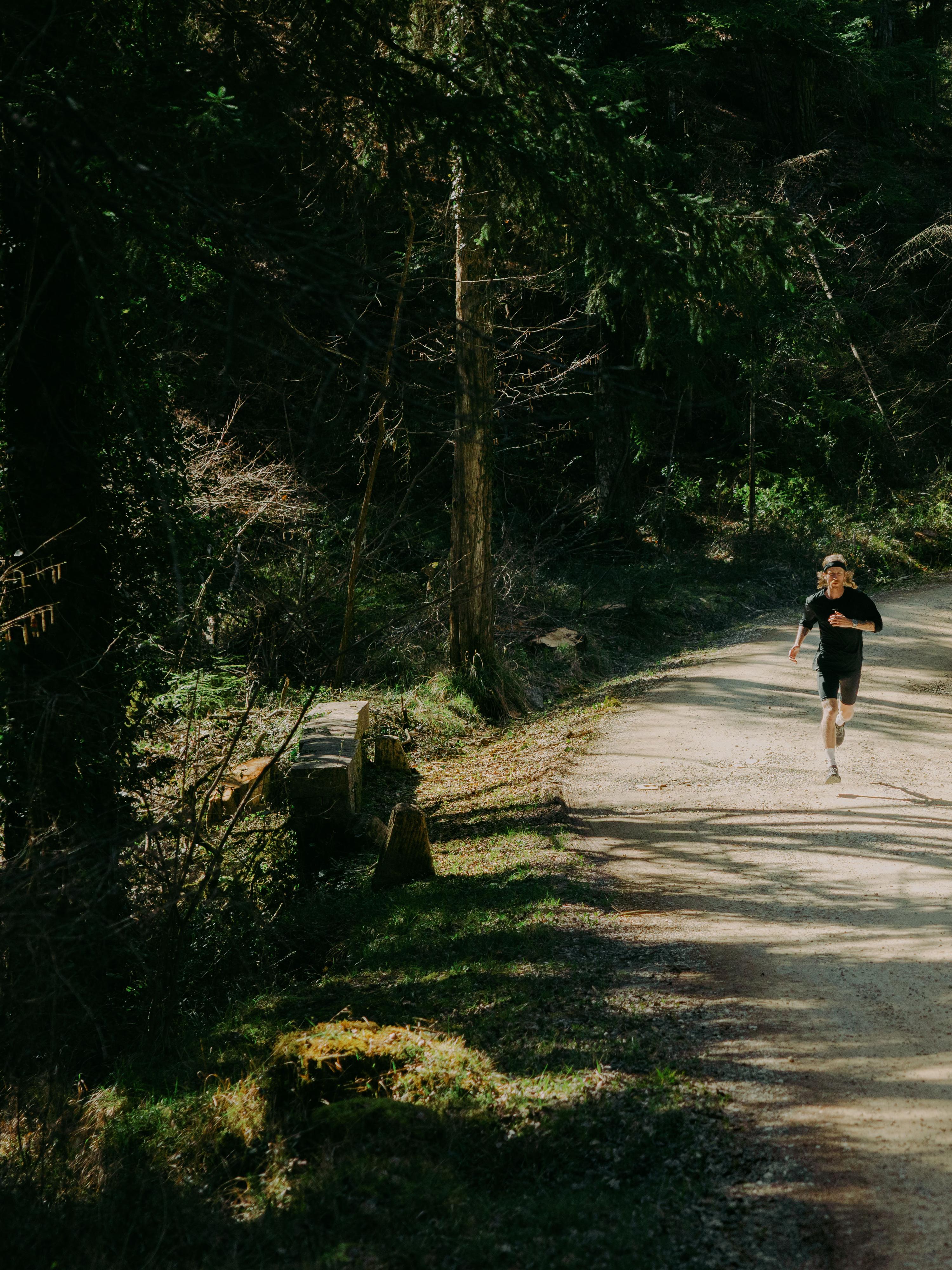 A person jogging on a sunlit, tree-lined dirt path in a forest, surrounded by lush greenery.