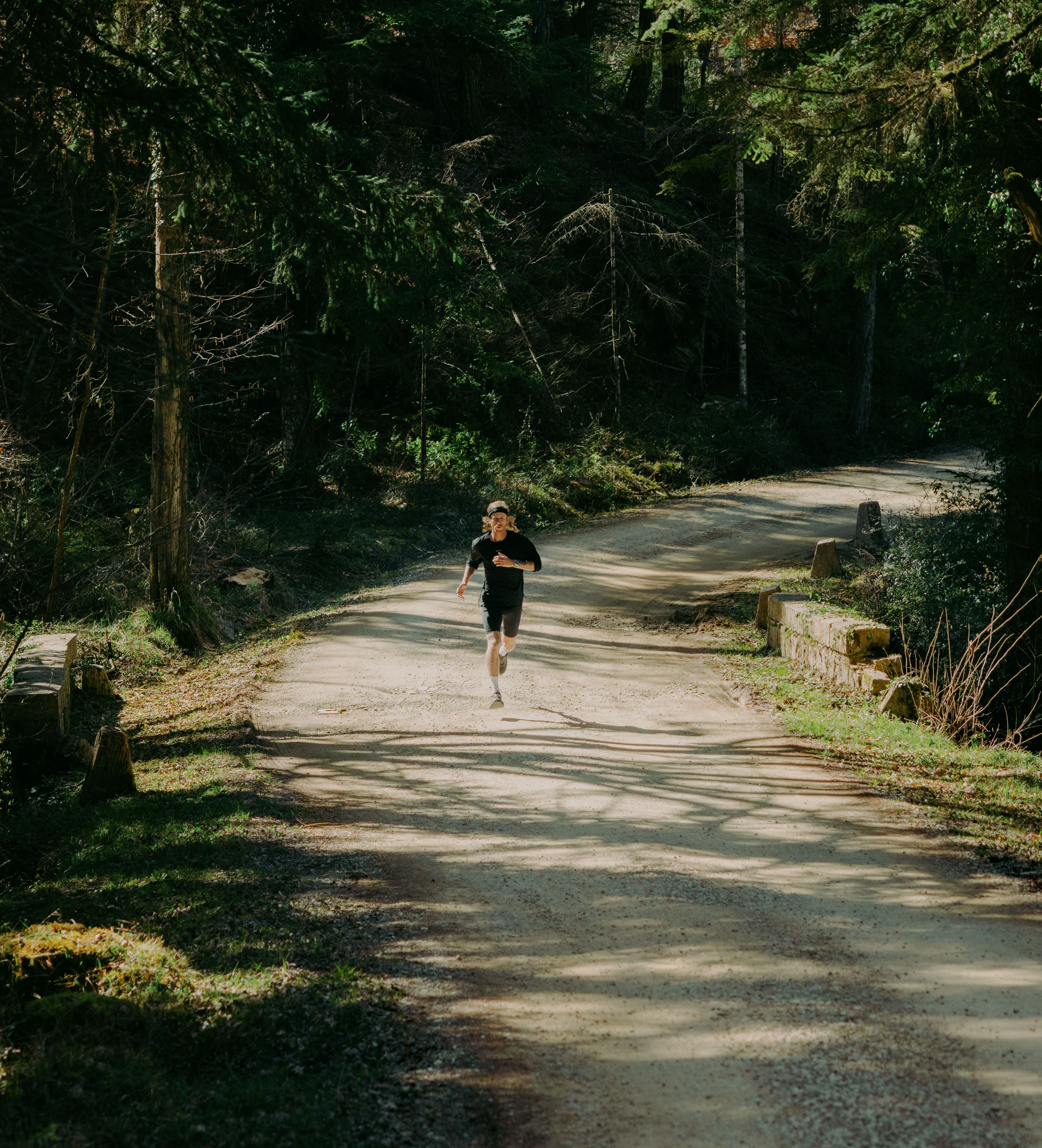 A person jogging on a sunlit, tree-lined dirt path in a forest, surrounded by lush greenery.