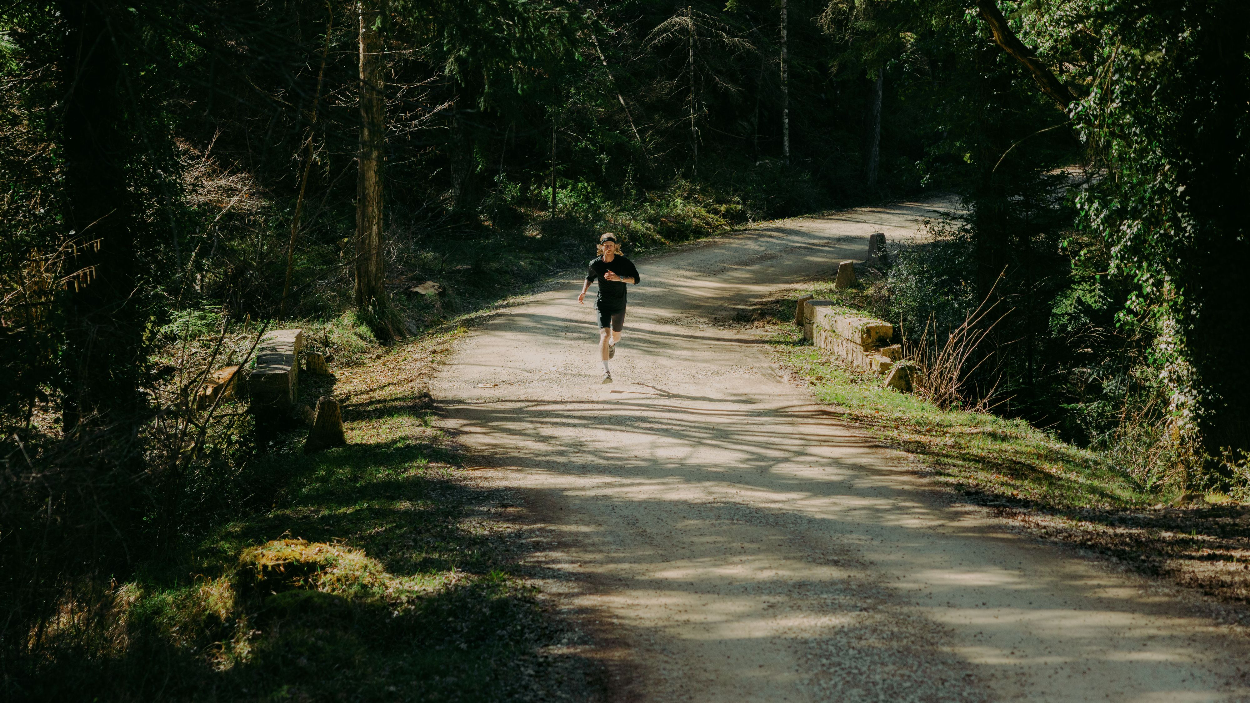 A person jogging on a sunlit, tree-lined dirt path in a forest, surrounded by lush greenery.