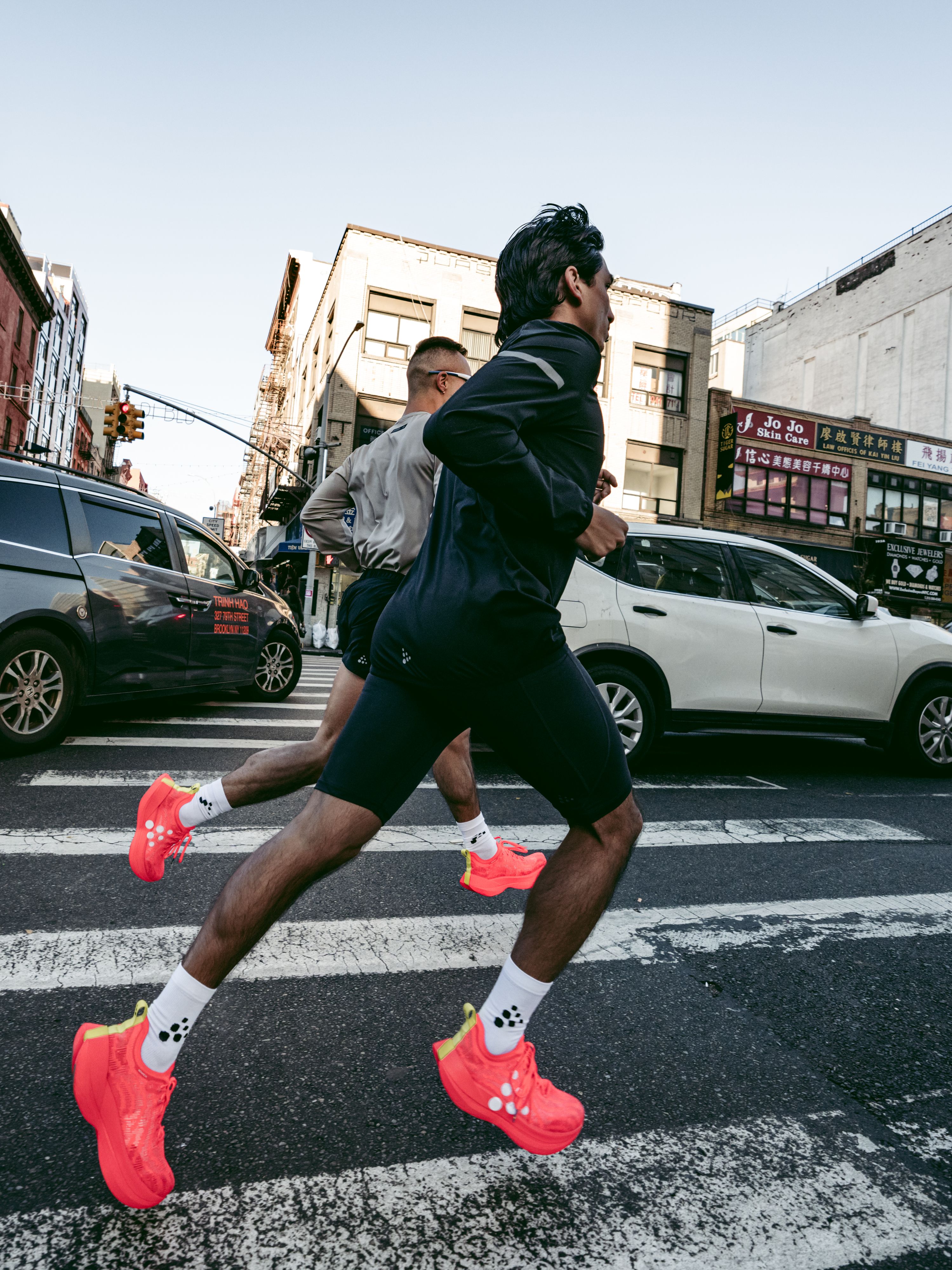 Two people jogging across a city crosswalk, wearing bright red shoes, with cars and buildings in the background.