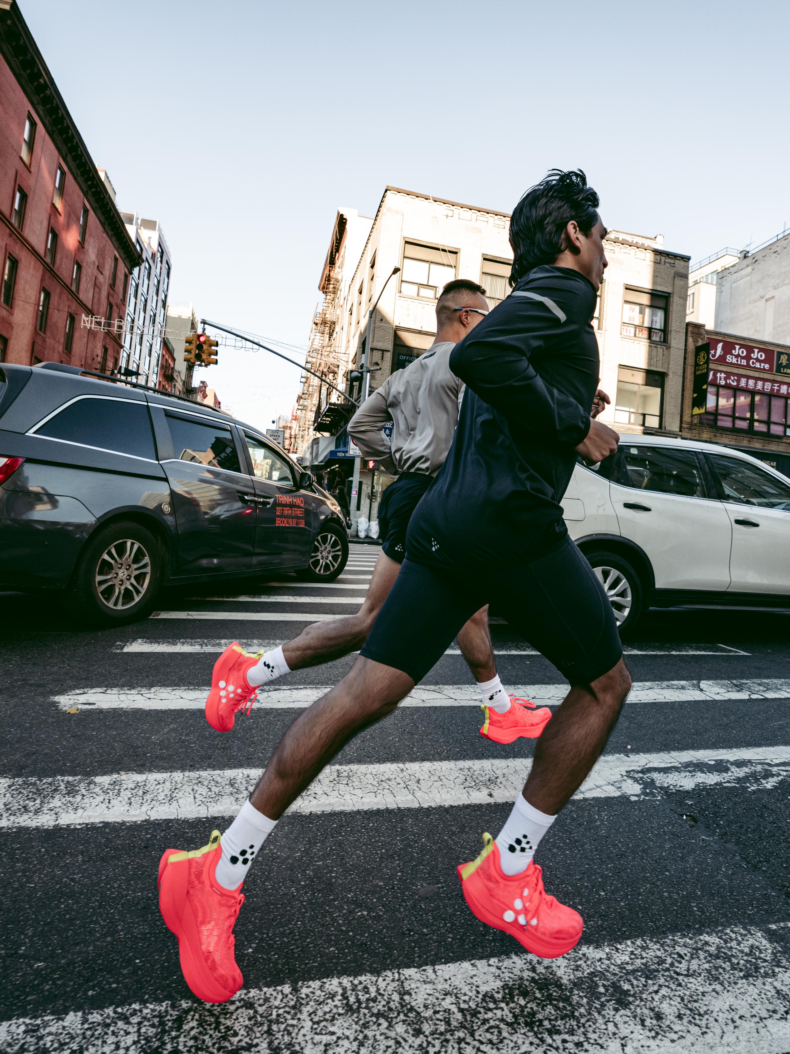 Two people jogging across a city crosswalk, wearing bright red shoes, with cars and buildings in the background.