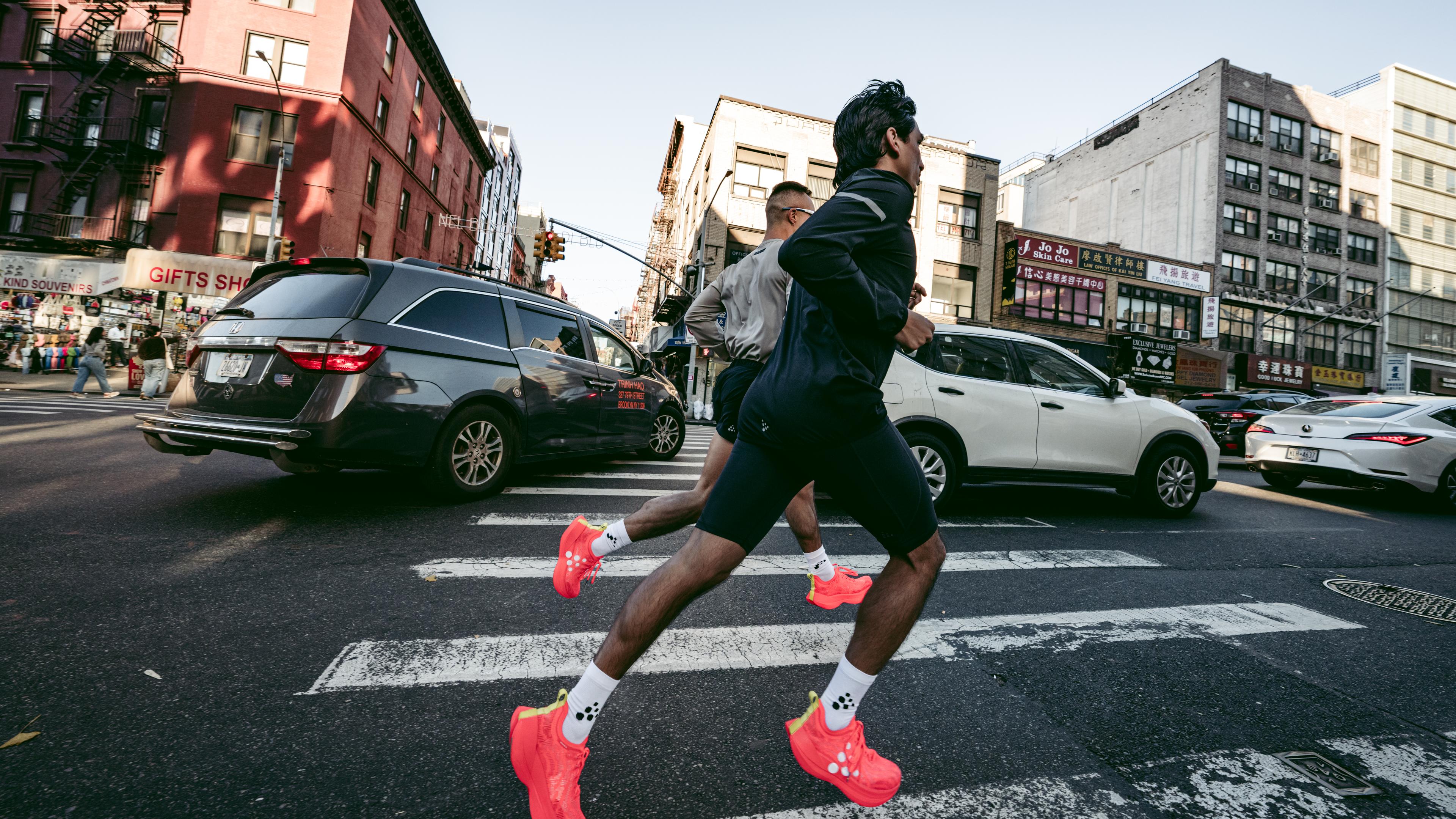 Two people jogging across a city crosswalk, wearing bright red shoes, with cars and buildings in the background.