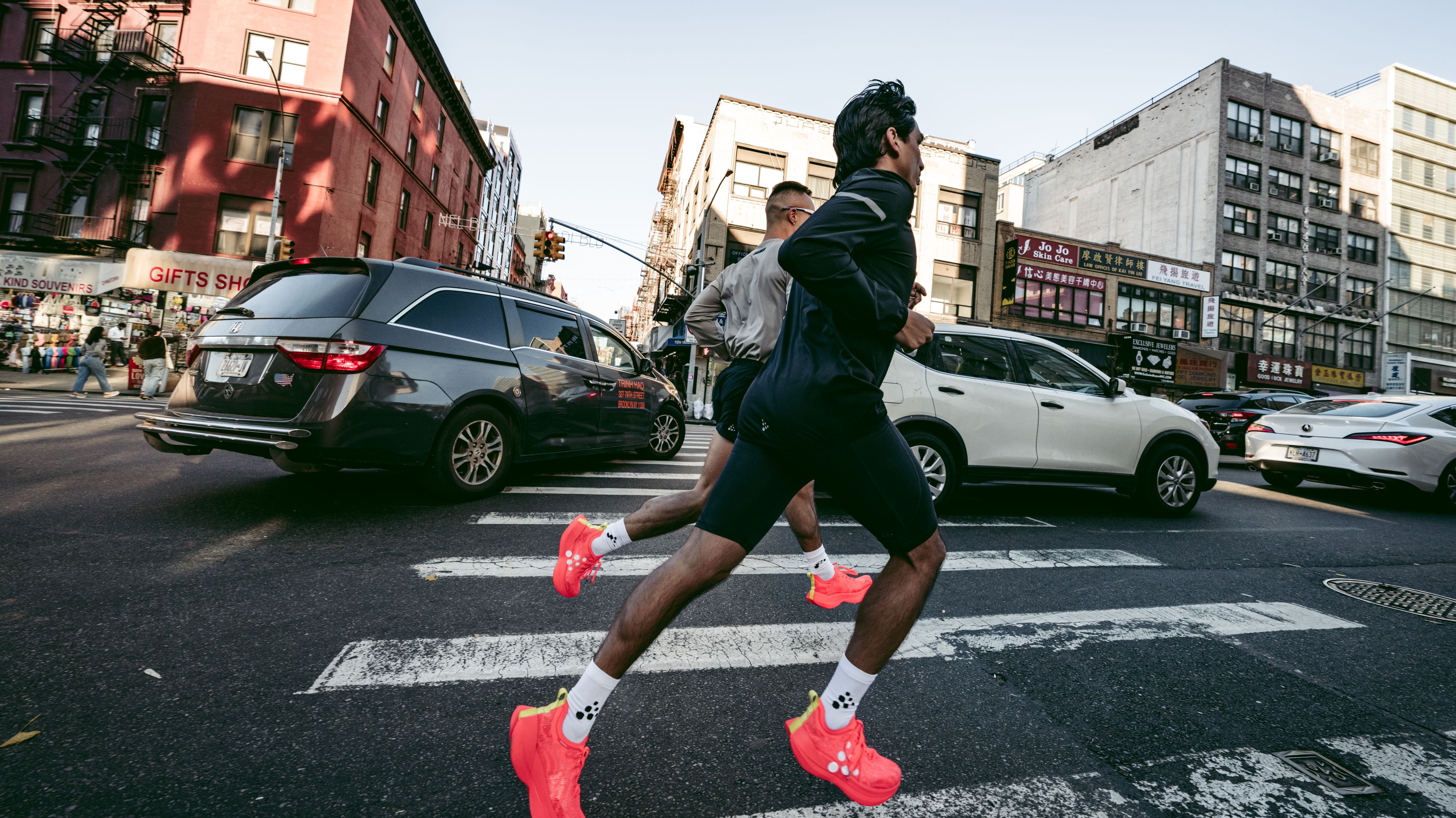 Two people jogging across a city crosswalk, wearing bright red shoes, with cars and buildings in the background.