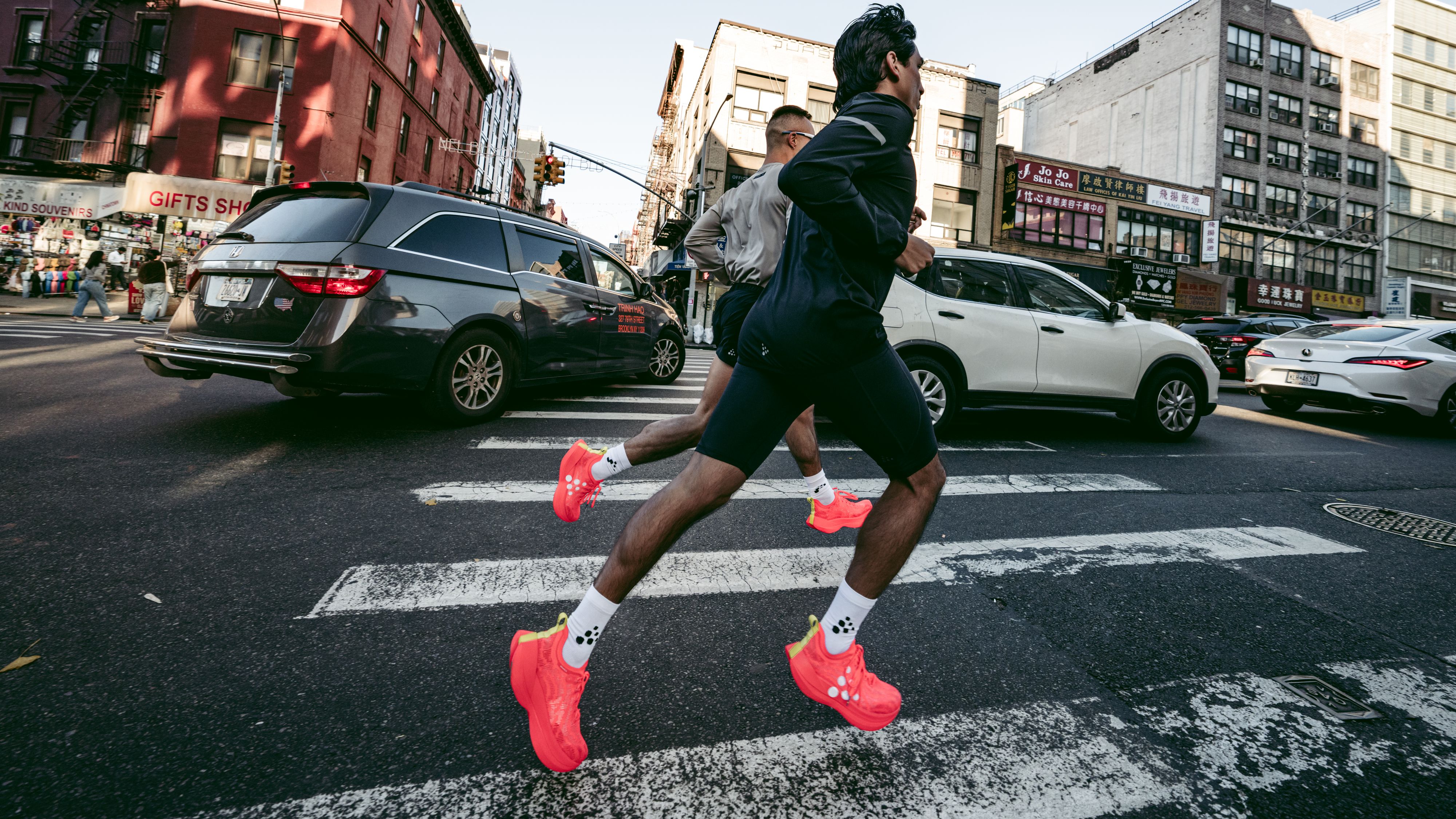 Two people jogging across a city crosswalk, wearing bright red shoes, with cars and buildings in the background.