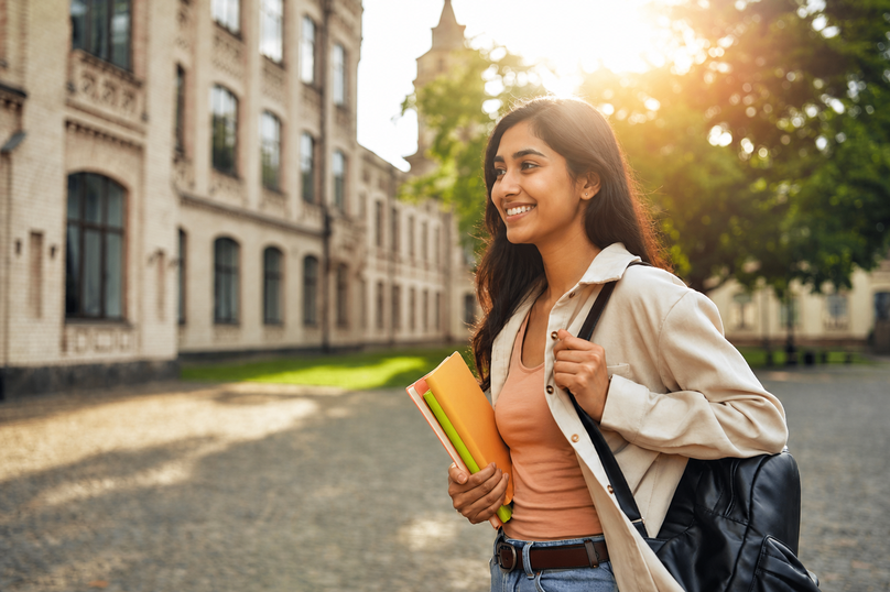A young woman student walking on campus with a backpack and books, enjoying the sunshine