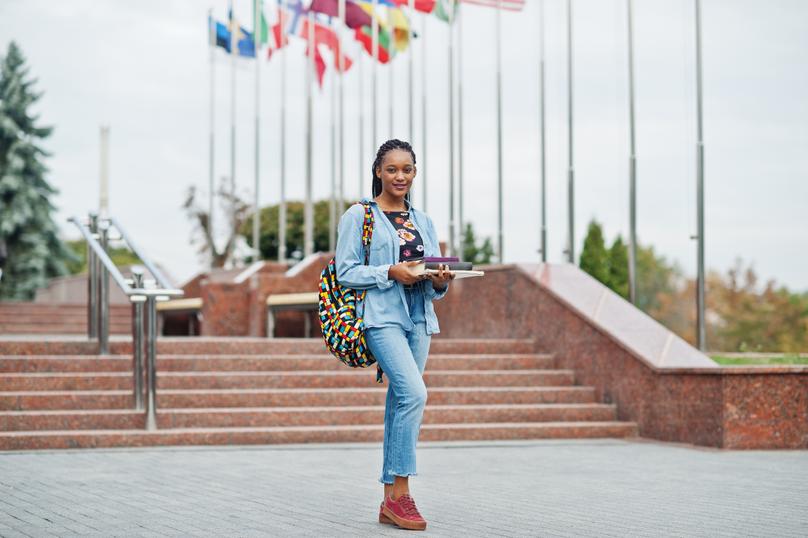 African student female posed with backpack and school items on yard of university, against flags of different countries.