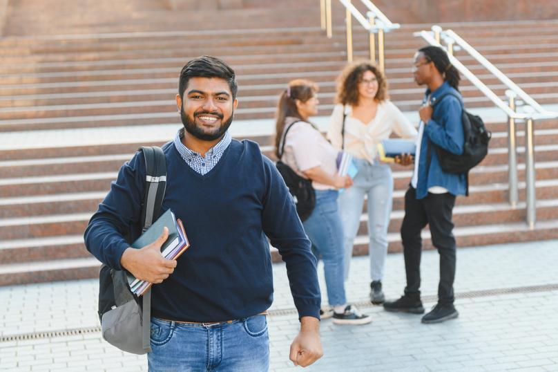 A young man with dark hair and a neatly trimmed beard smiles at the camera. He wears a blue sweater over a patterned collared shirt and blue jeans. He carries books and has a grey backpack slung over his shoulder. In the background, three other people can be seen talking near a stairway. This cheerful image is perfect for representing campus life, education, friendship, and the excitement of learning. The subjects and setting offer strong visual storytelling for advertising, academic materials, or motivational content.
