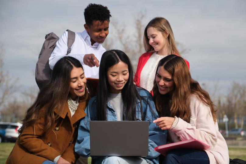 Happy multi ethnic students working together on a project using a laptop computer outdoors in a university campus