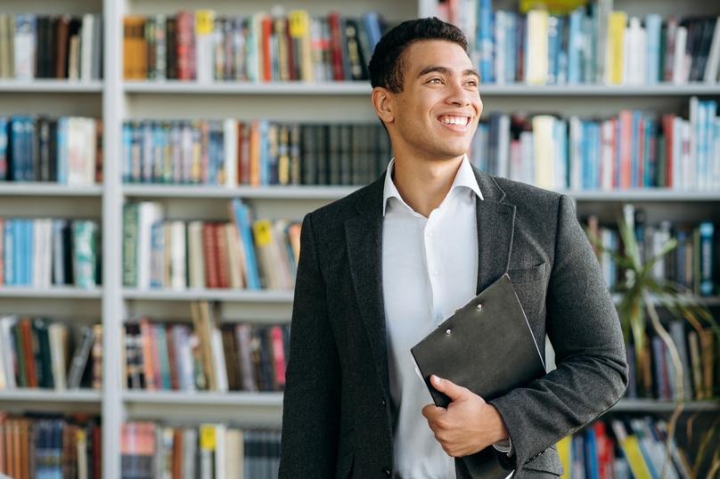A smiling young adult man wearing a gray jacket and white shirt is visible from the waist up in front of a large bookshelf. He is holding a dark clip board in one hand. The photo has clear lighting and would be appropriate for business and office related creative projects.