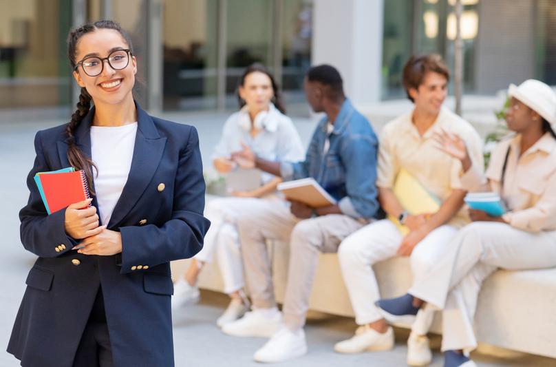 Photo of cheerful beautiful young woman in formal outwear and eyeglasses posing over multicultural group of students resting at university campus