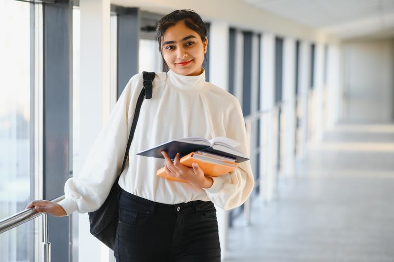 A young woman stands smiling in a bright hallway in an urban school building. She carries books and a backpack, and wears a white top and black pants. Light streams in through the windows.